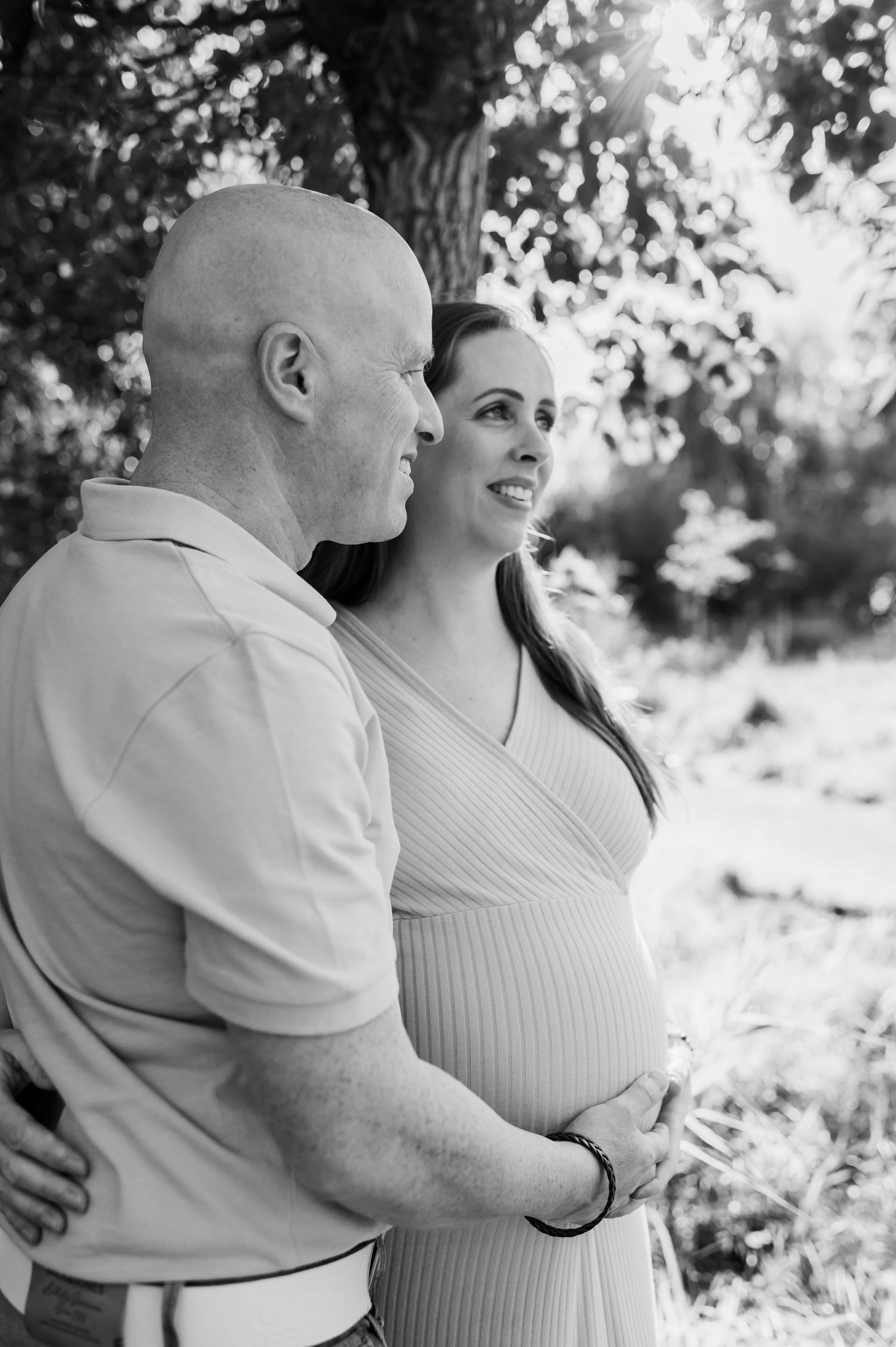 Black and white photo of expectant couple embracing outdoors under trees.