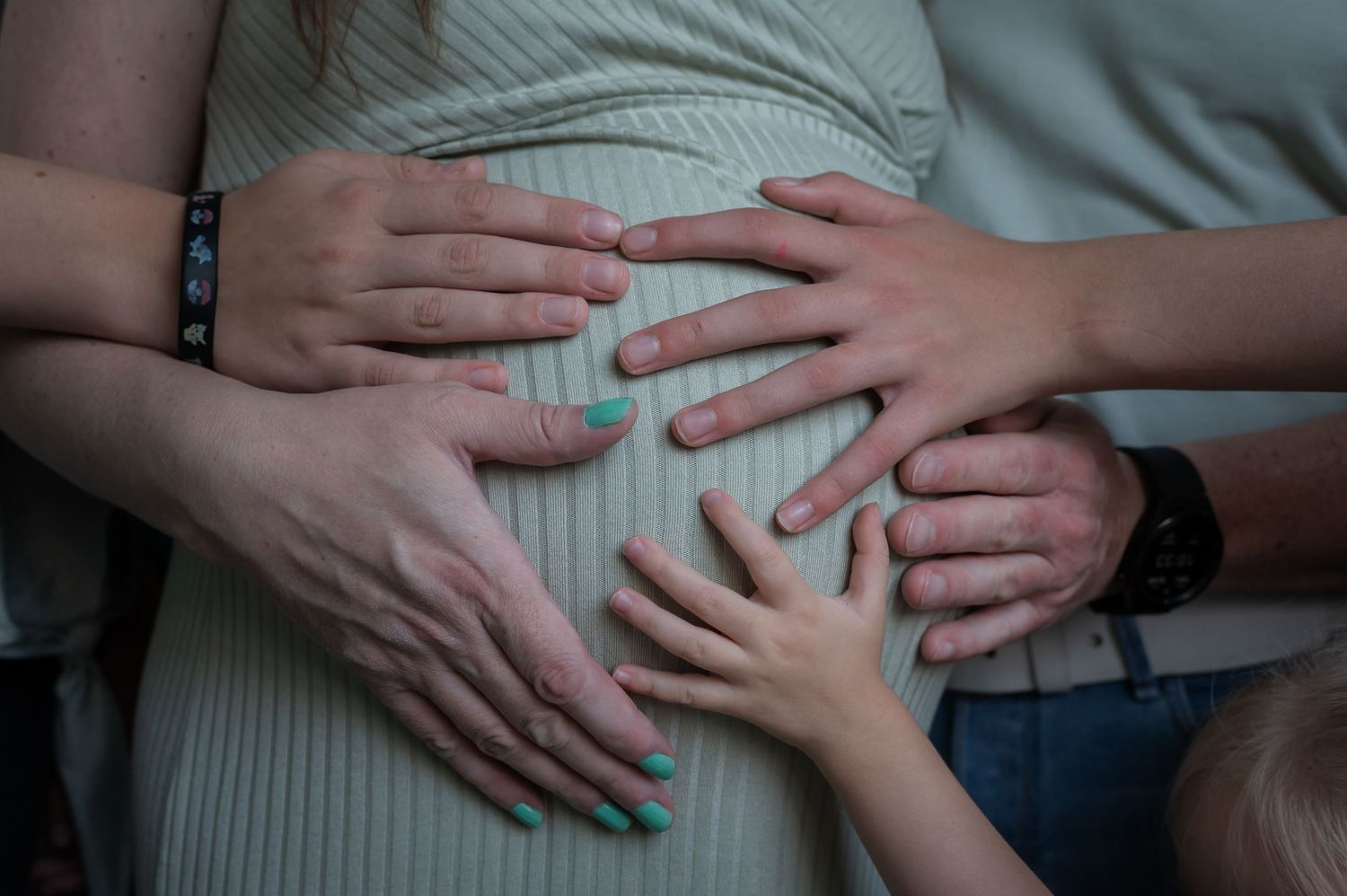 Multiple hands resting on a pregnant woman's belly, including a child's hand with teal nail polish.