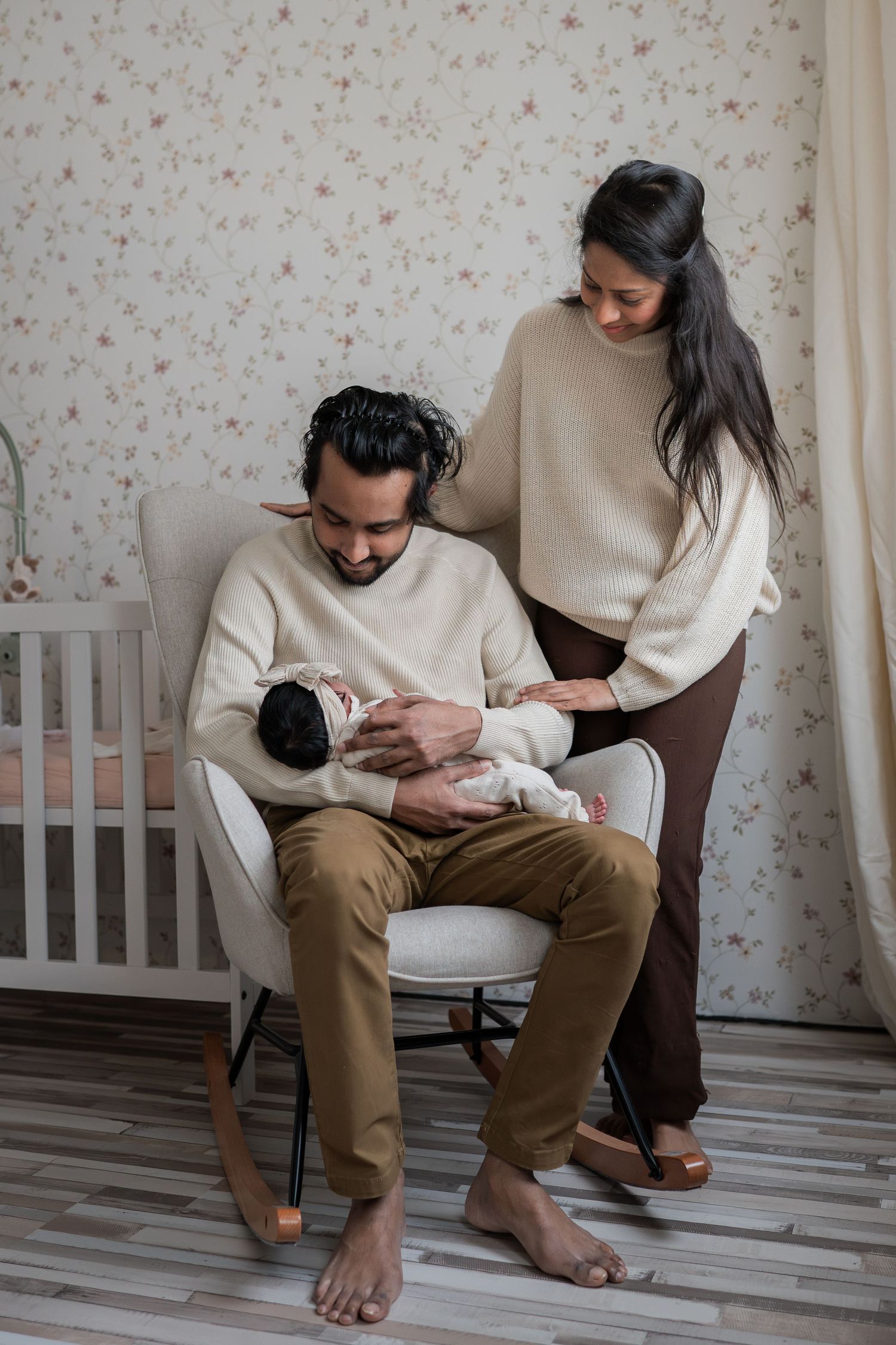 A couple in neutral sweaters admires their newborn baby in a cozy nursery with floral wallpaper and a white crib.