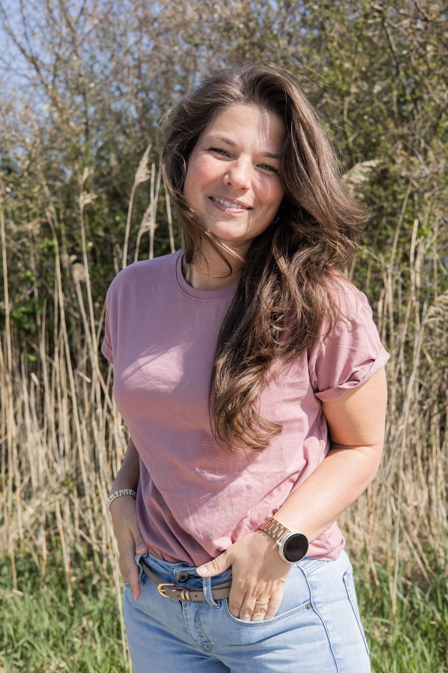 Smiling young woman in pink t-shirt and jeans posing outdoors in a sunny natural setting with tall grass. jouw fotograaf Lelystad, Almere,