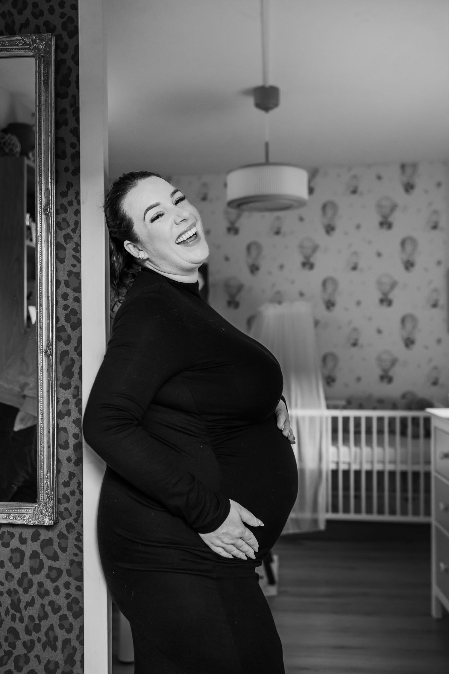 Black and white portrait of laughing pregnant woman in black dress cradling baby bump near nursery crib.