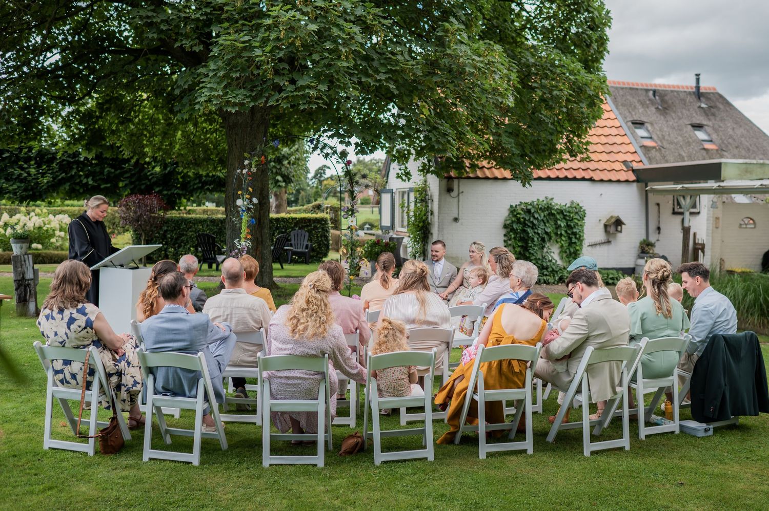 Outdoor garden party with guests seated at round tables under trees near a white building.