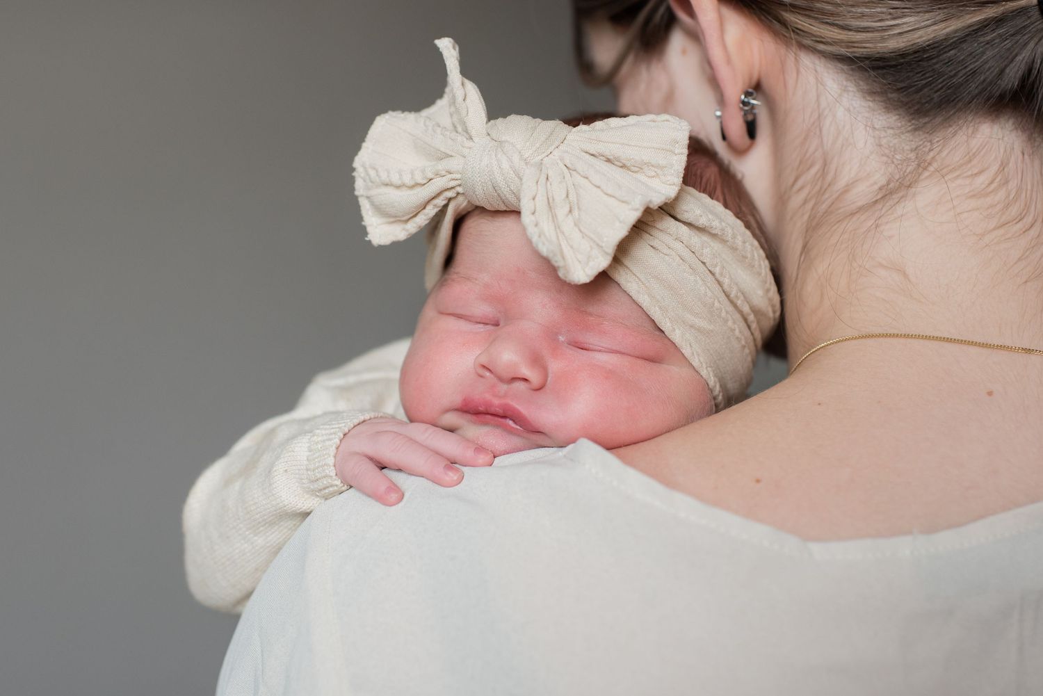 Newborn baby sleeping peacefully on mother's shoulder, wearing a cream bow headband and white outfit.