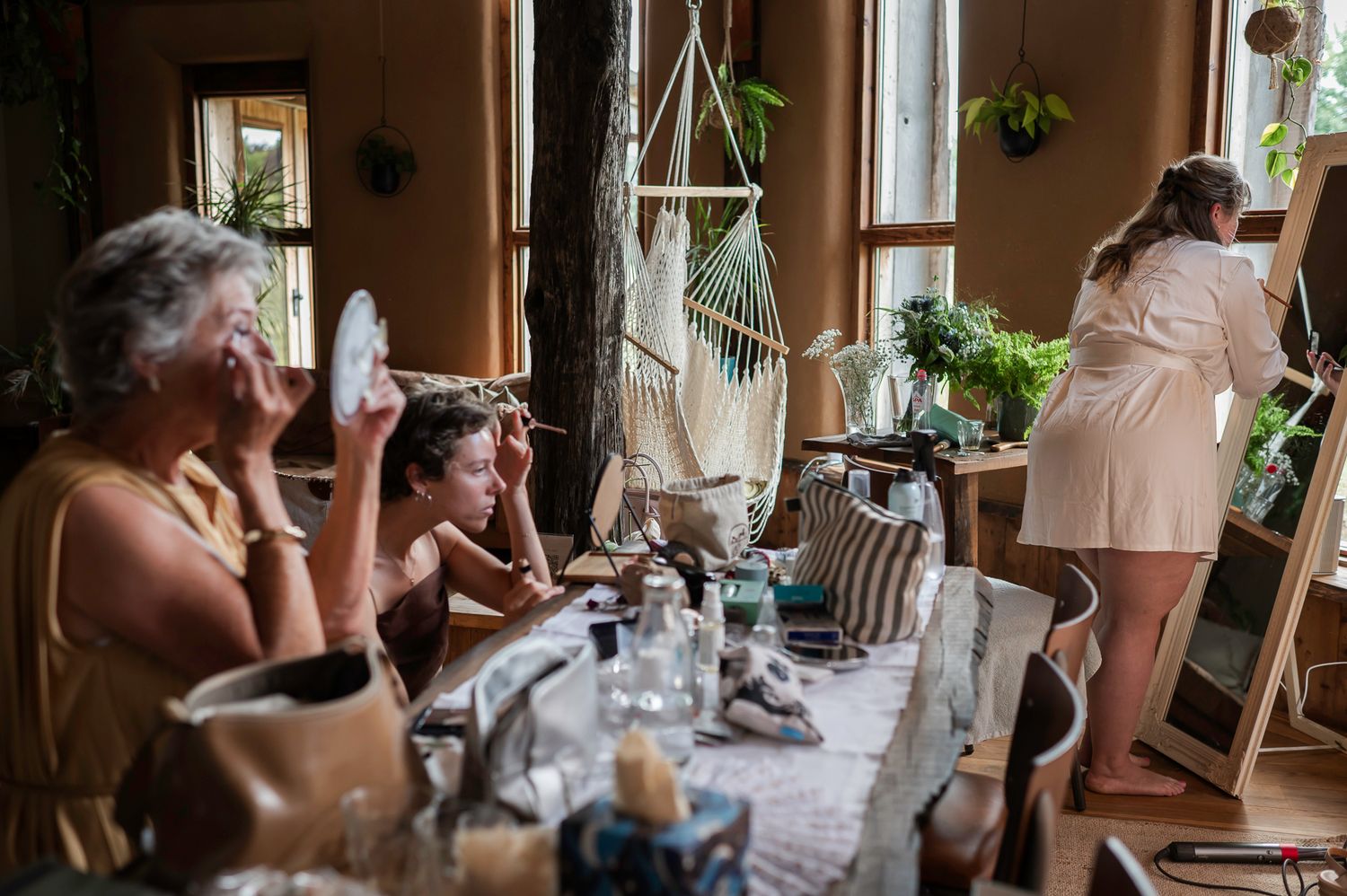 Women getting ready in a bohemian room with hanging chair, plants, and vanity table with makeup.