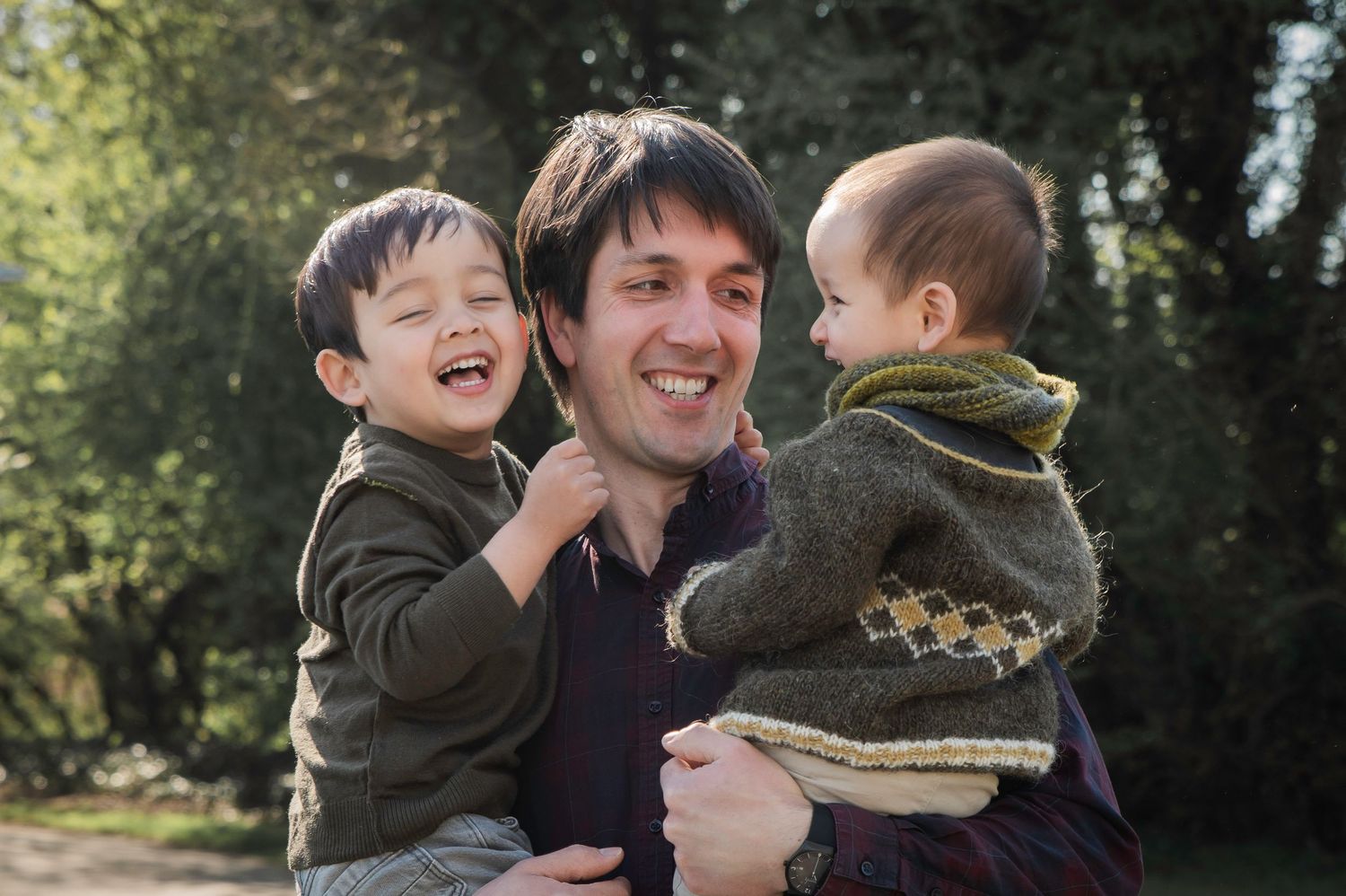 A smiling father holds two young children outdoors, surrounded by lush green trees on a sunny day.