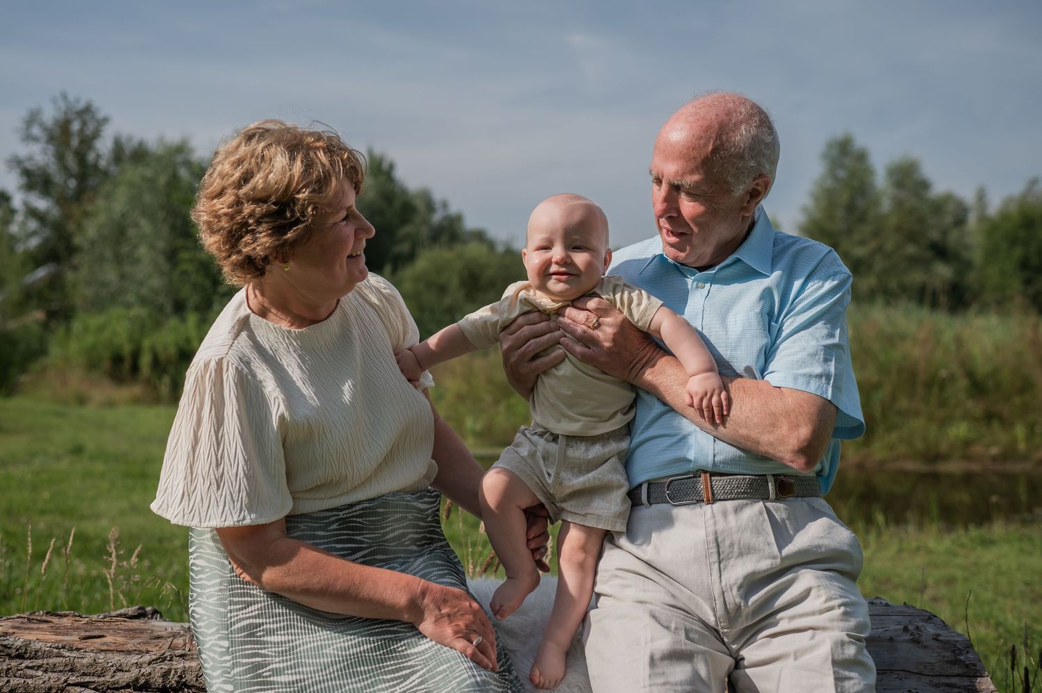 Grandparents sitting outdoors on a log, smiling and holding a happy baby in a green nature setting.
