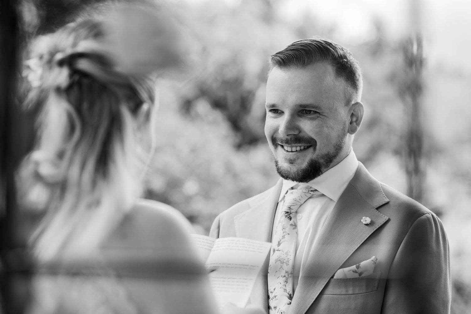 Black and white photo of a smiling groom in a suit looking lovingly at his bride during a wedding ceremony.