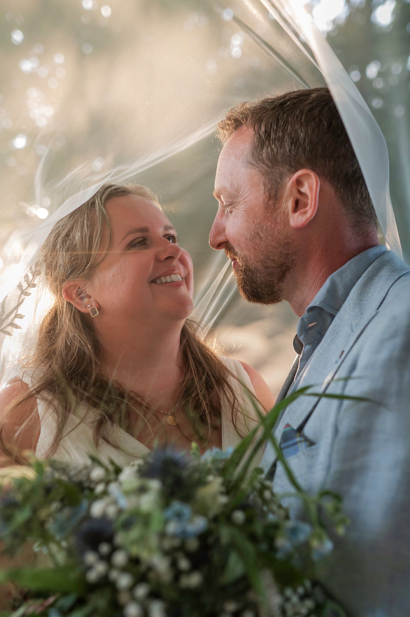 A bride and groom share a romantic moment under a wedding veil, surrounded by soft golden light and a bouquet of wildflowers.