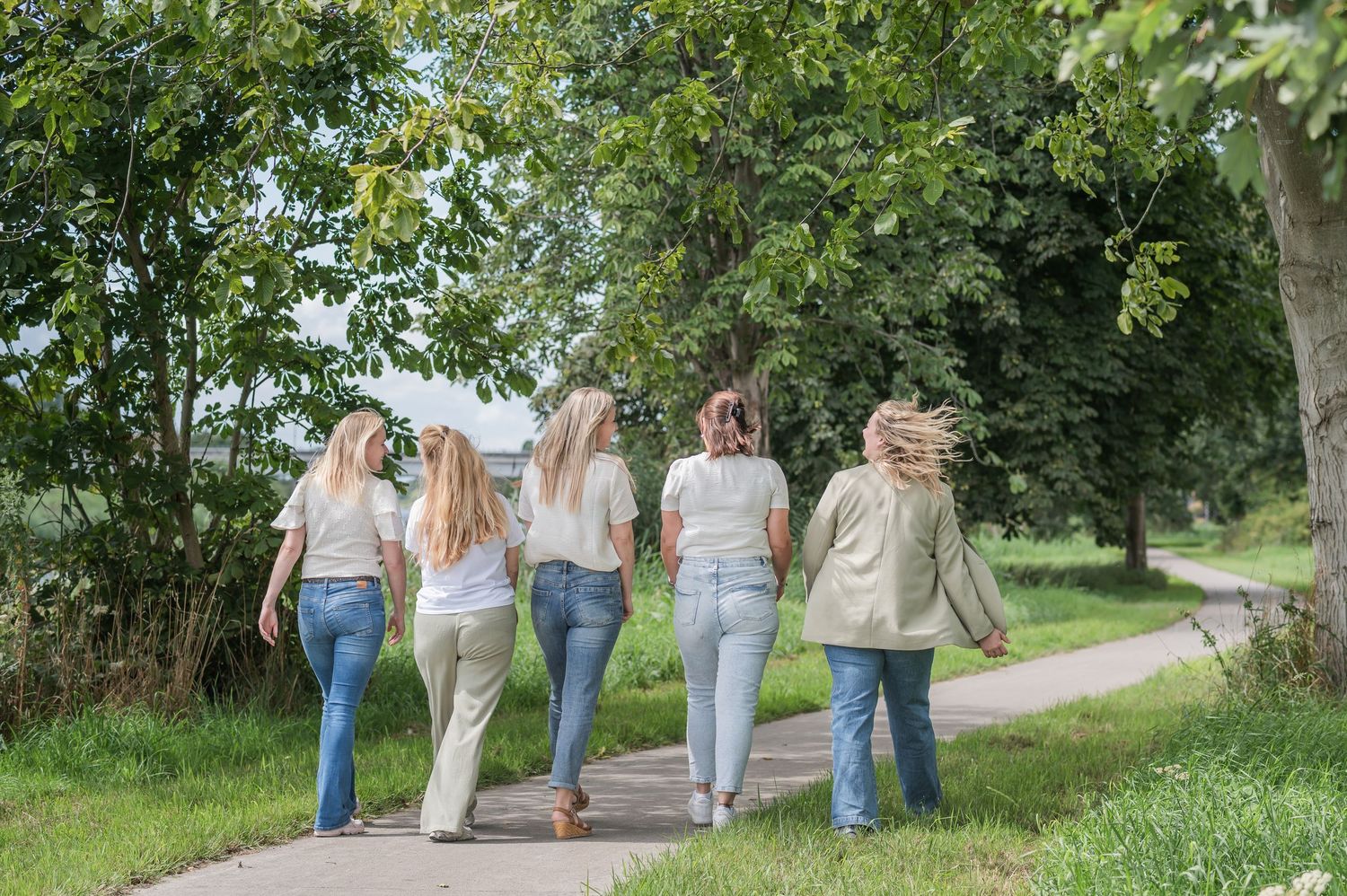 Five women walking together on a scenic tree-lined path surrounded by lush green grass and foliage.