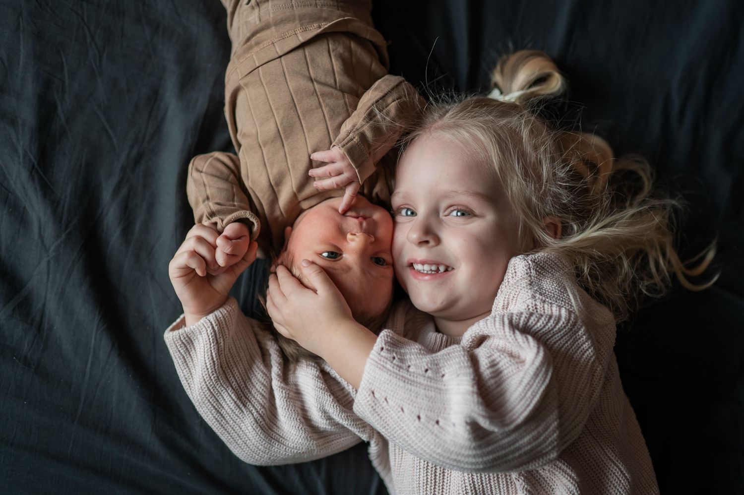 A smiling blonde toddler girl poses with a newborn baby on a dark blanket. Lelystad, Almere, Dronten, Flevoland