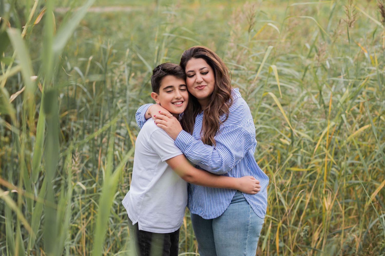 A woman and boy share a warm hug in a lush green field, smiling together in a heartfelt outdoor portrait.