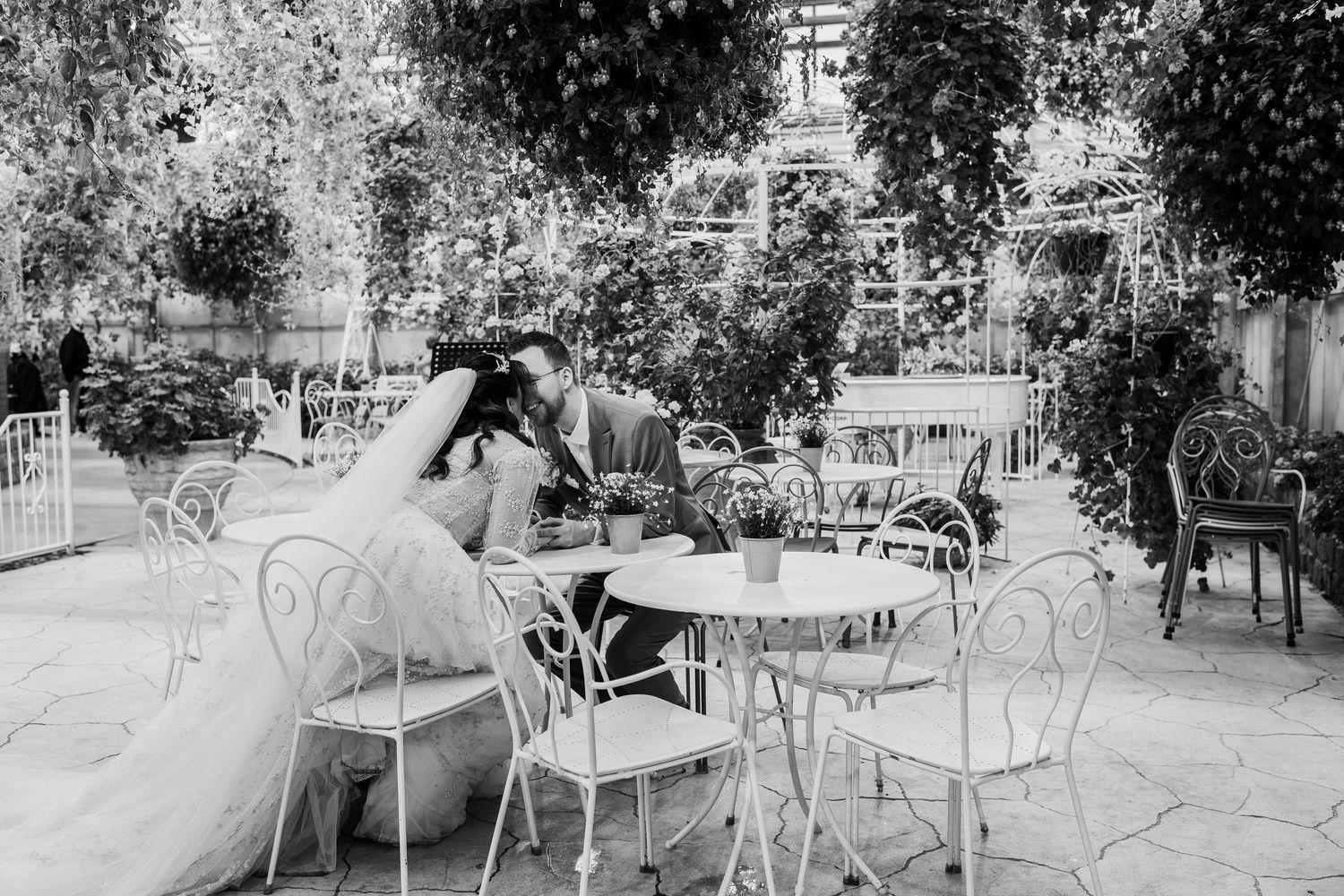 Black and white photo of a bride and groom sharing an intimate moment at a garden cafe with ornate iron chairs.