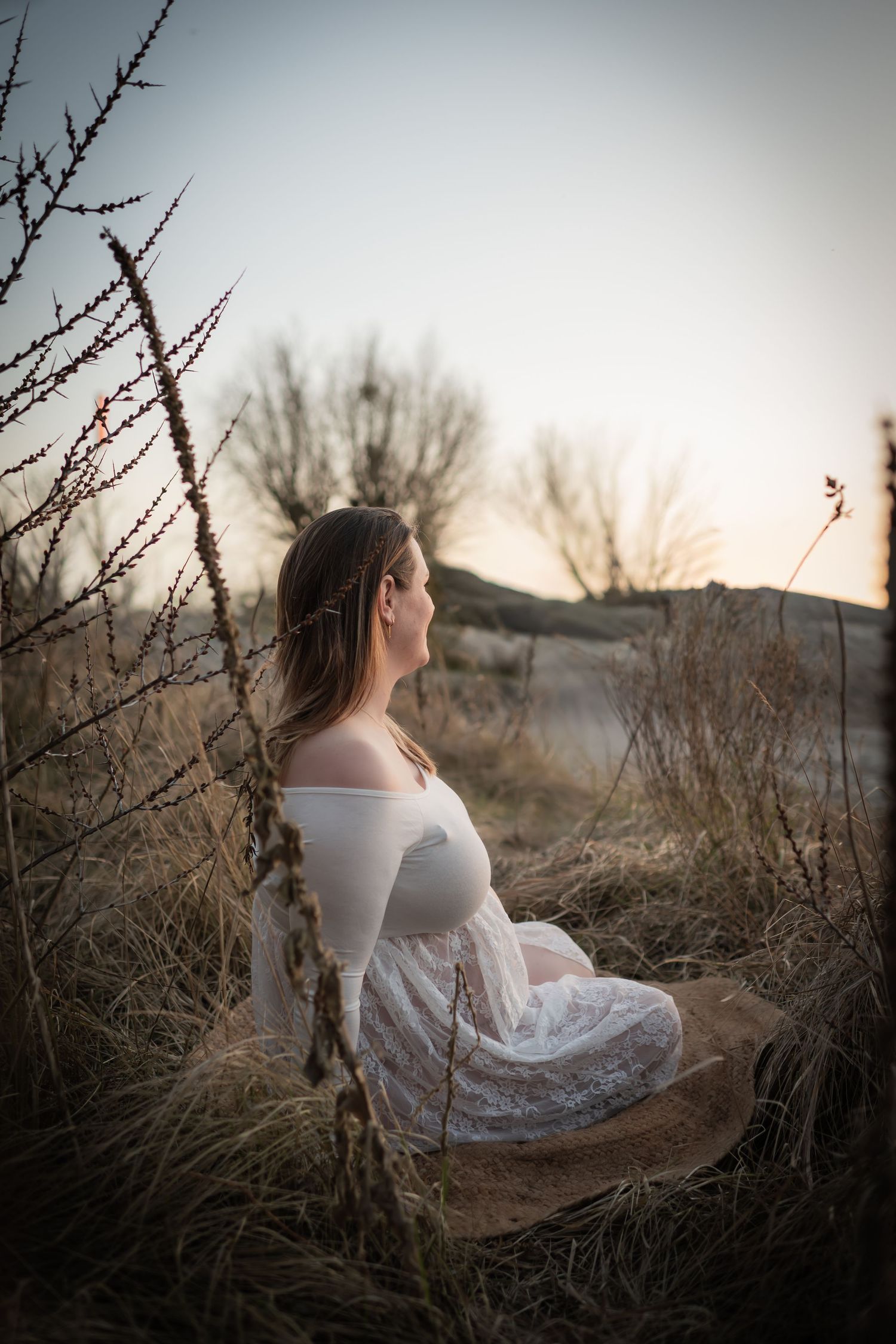 Pregnant woman in white lace dress sitting peacefully in a field at dusk.