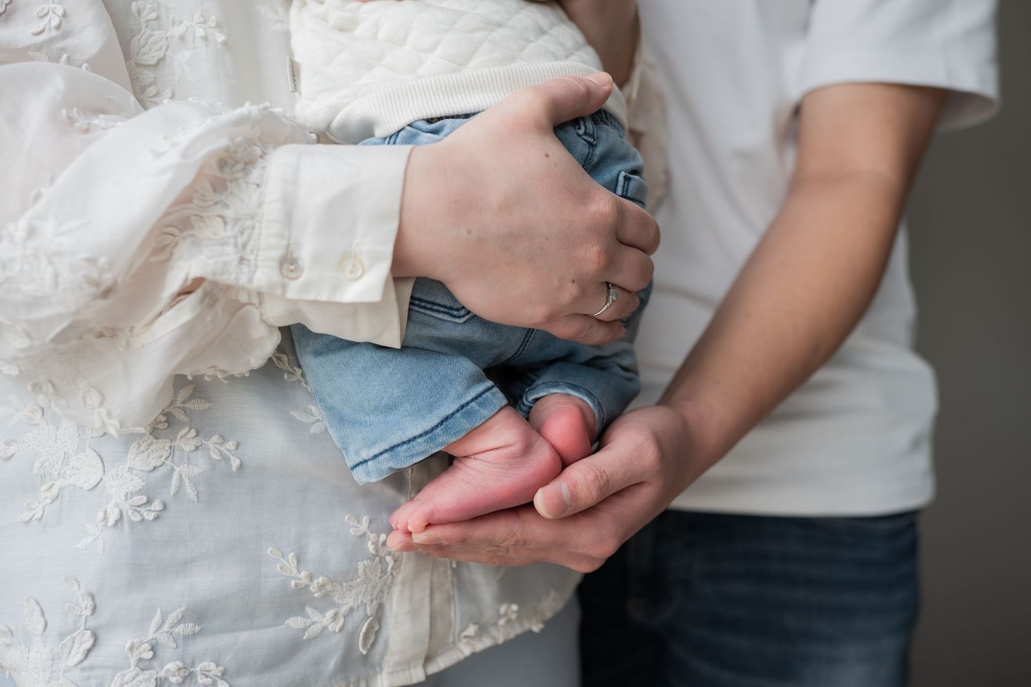 Couple tenderly holding newborn baby hands, mother in white lace dress and father in white shirt, close-up maternity photo.