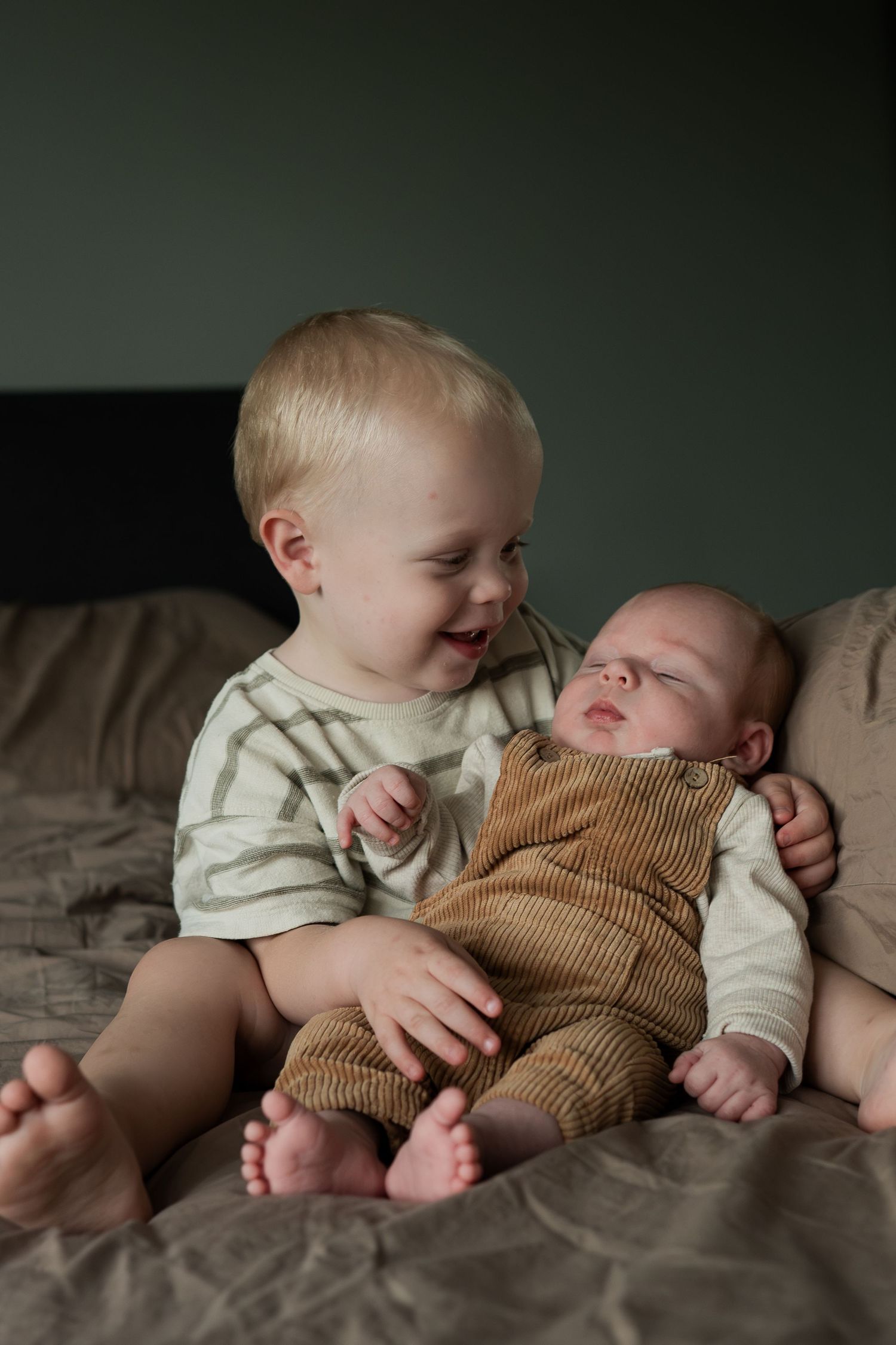 Toddler smiling while holding a sleeping newborn baby in a brown corduroy outfit on a bed.