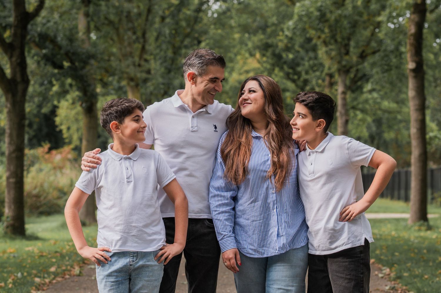 Happy family of four with two teenage boys walking together in a tree-lined park path.