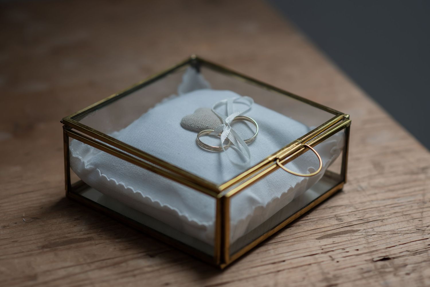 Wedding rings displayed on white pillow inside elegant gold-framed glass box on wooden surface.