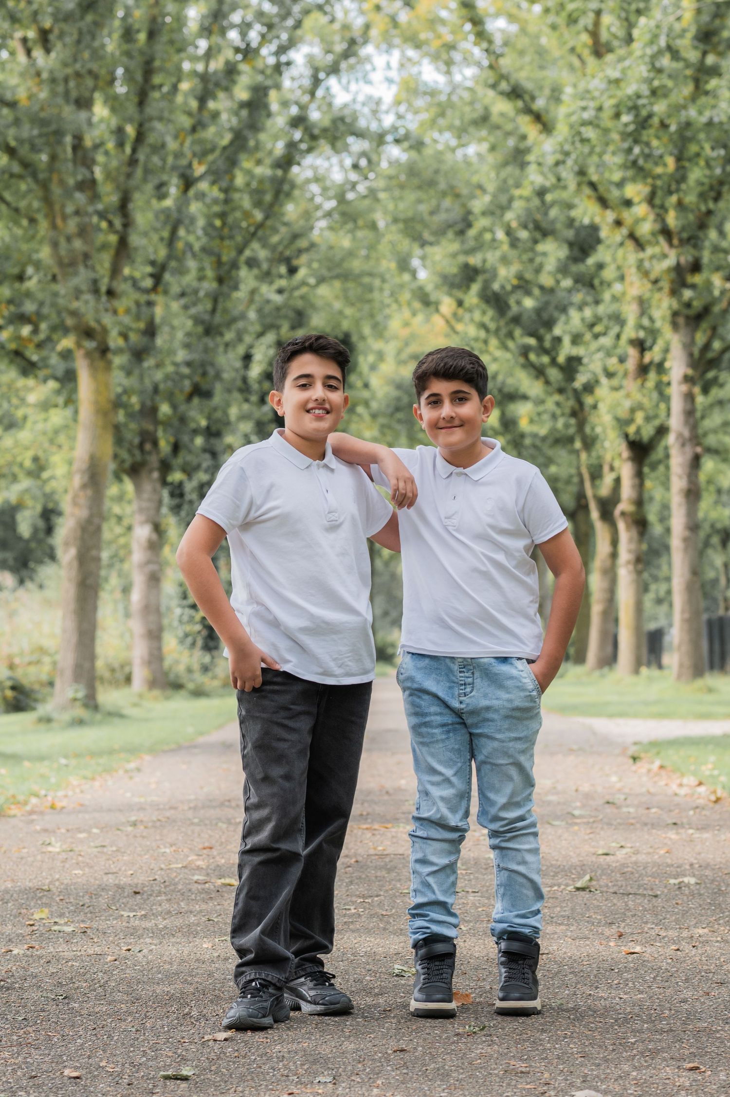 Two young boys in white polo shirts pose together on a tree-lined park path, smiling at the camera.