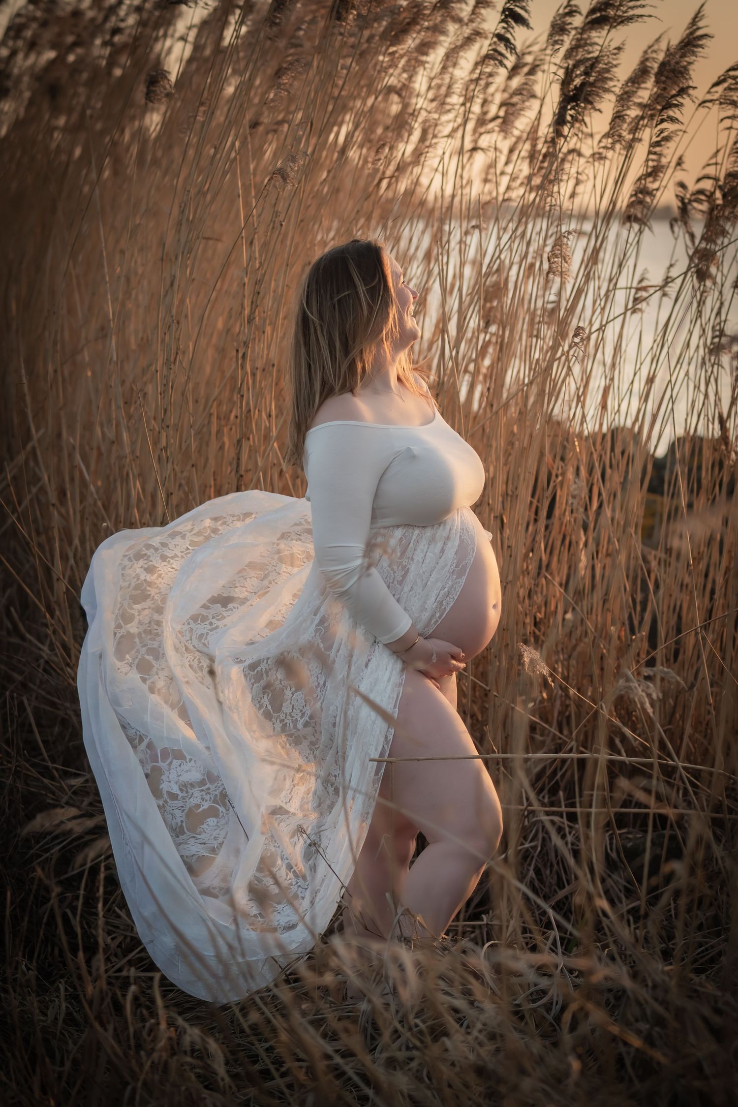 Pregnant woman in white lace dress standing among golden reeds at sunset.