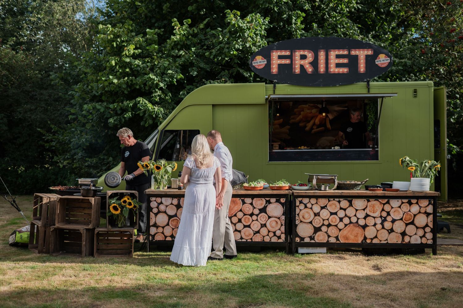 Green food truck labeled FRIET serving customers outdoors, decorated with sunflowers and firewood in a lush park setting.