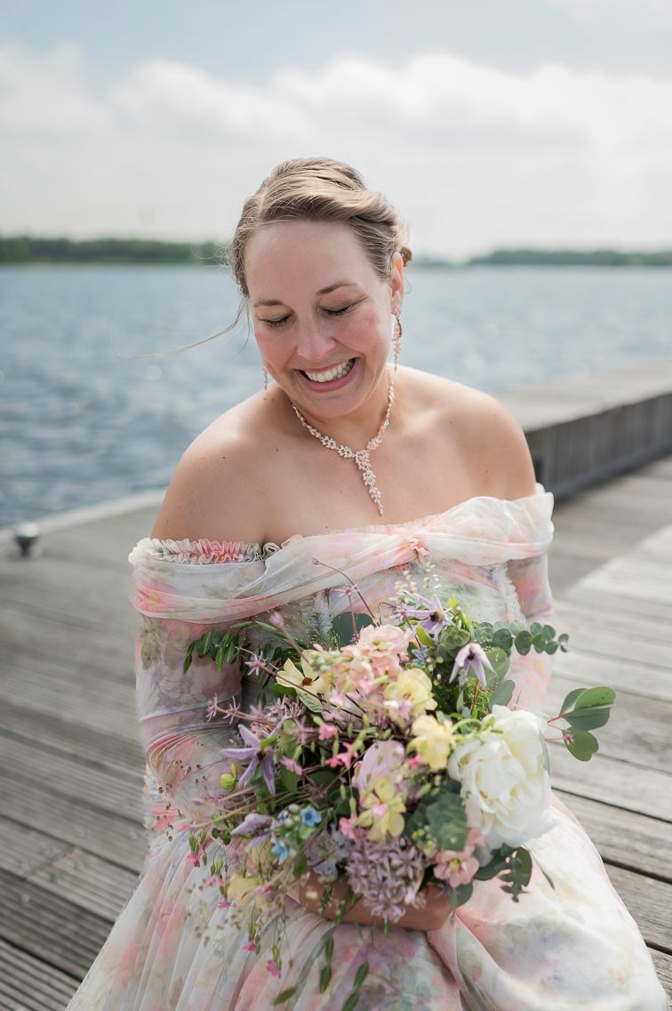 Smiling bride in floral off-shoulder gown holds colorful bouquet on wooden dock with lake backdrop.