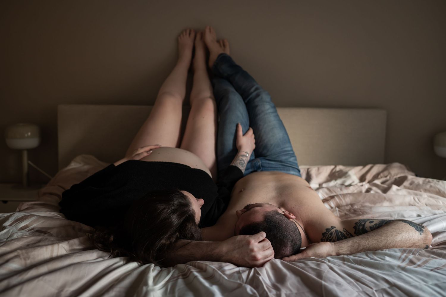Couple lying together on a bed in an intimate embrace, wearing casual clothing in a softly lit bedroom.