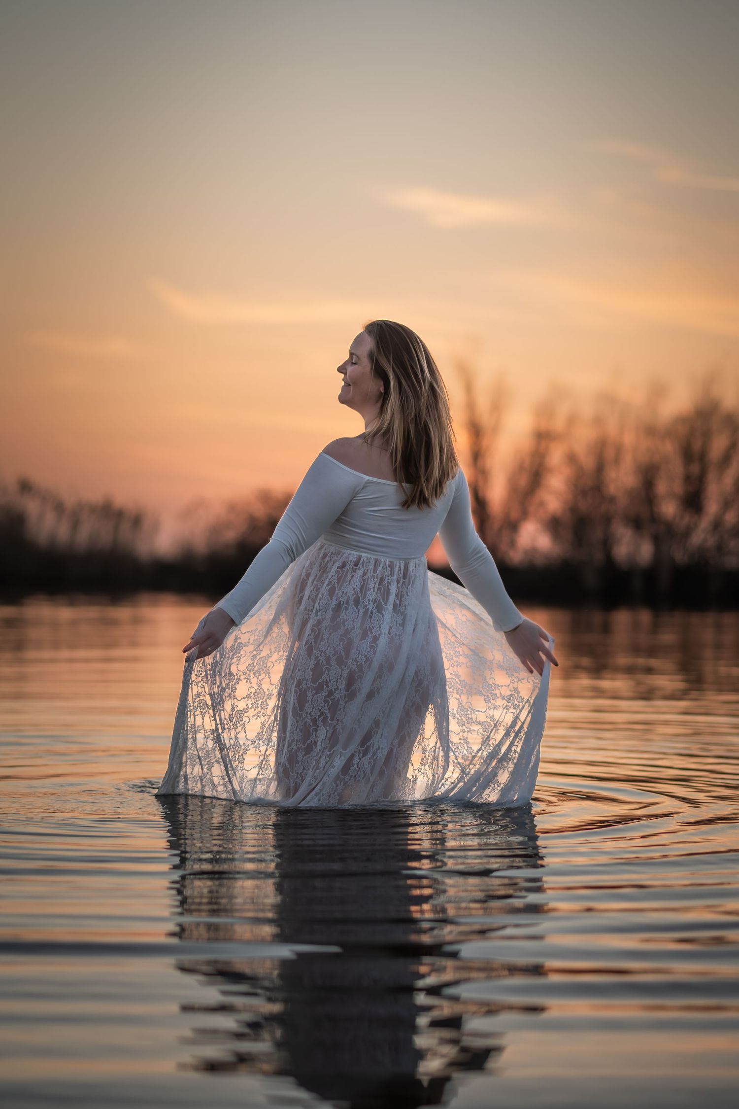 Woman in white lace dress standing in calm water at sunset, with warm golden sky and silhouetted trees in background.
