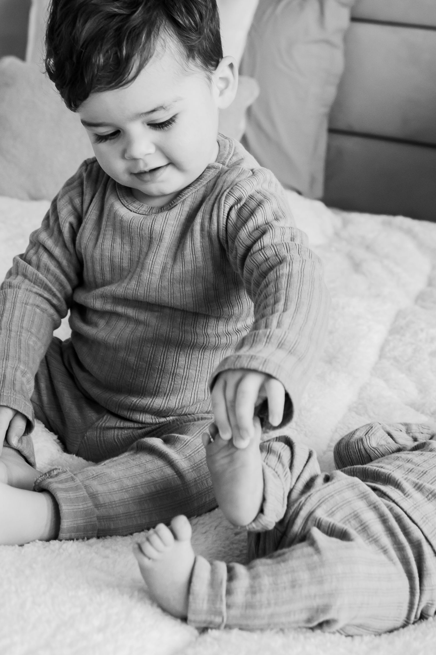 Adorable toddler in ribbed pajamas sitting on a white bed playing with their feet in black and white.