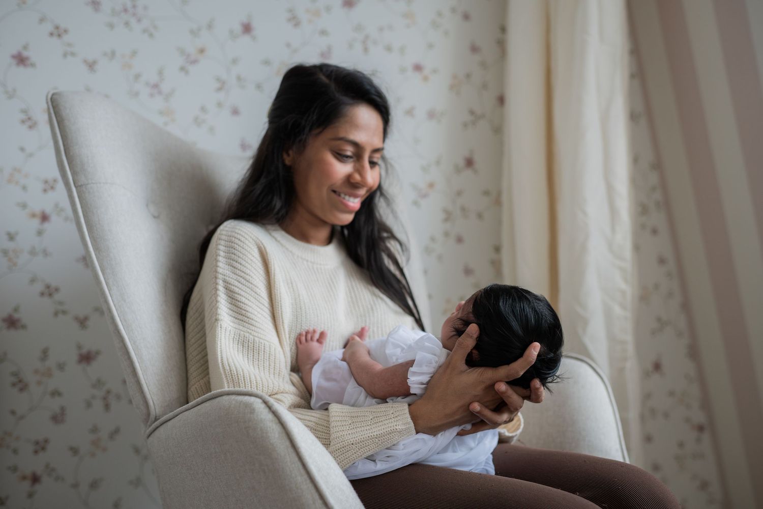 A smiling mother in a cream sweater holds her newborn baby while sitting in a cozy armchair. Lelystad, Almere, Dronten, Flevoland