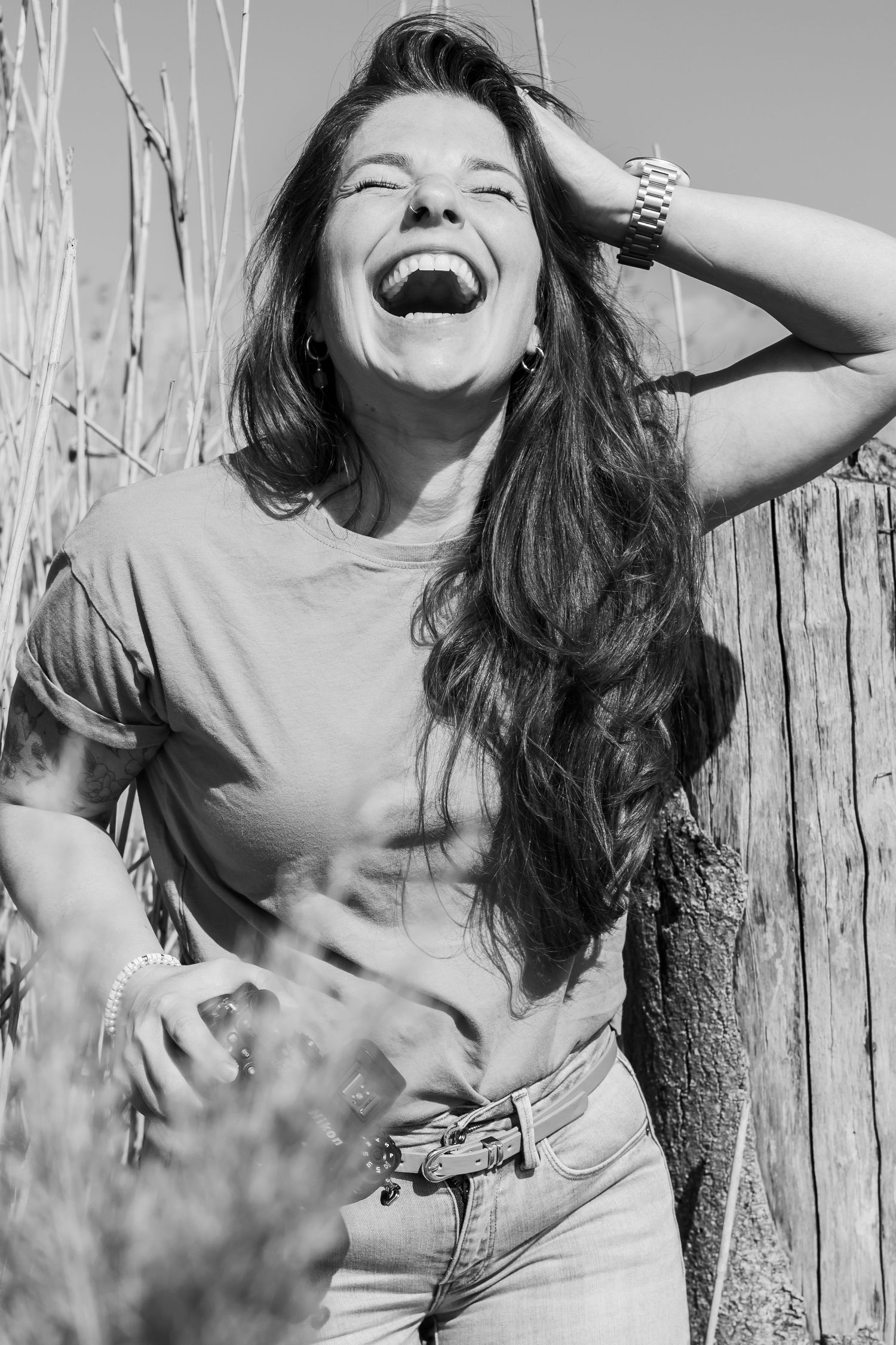 Black and white photo of a laughing woman with long hair, leaning against a fence outdoors. Jouw fotograaf Lelystad, Almere, Dronten