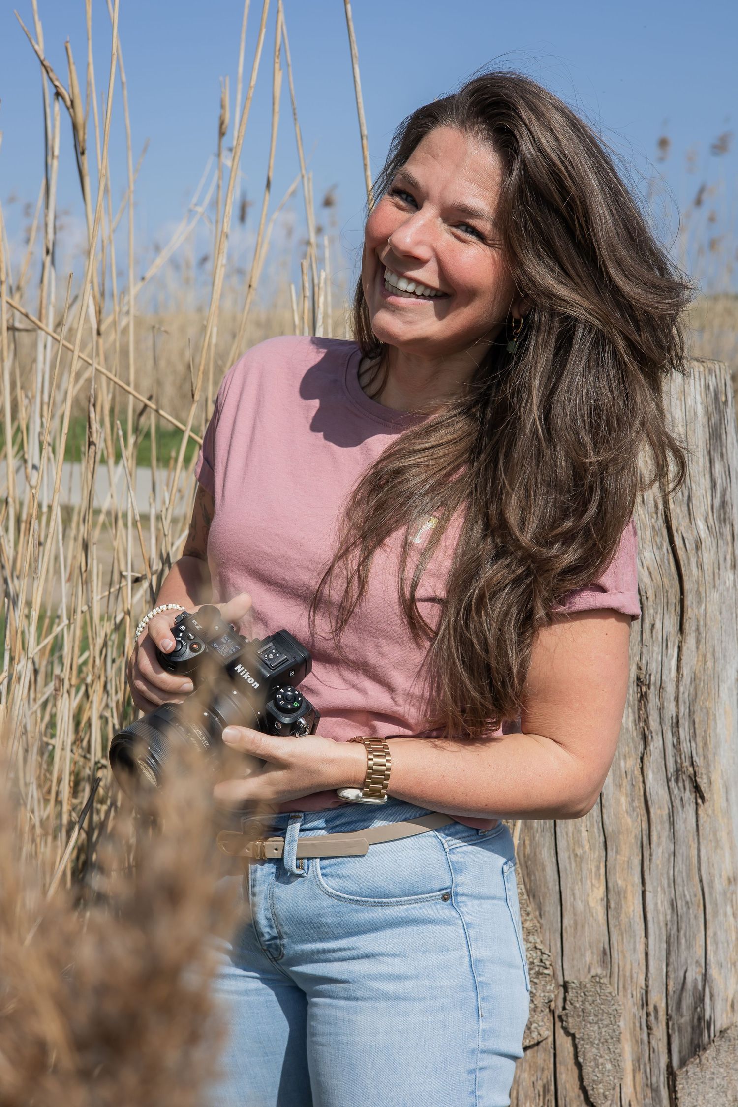 Smiling brunette woman in pink shirt and jeans holding a small dog outdoors near wooden fence and tall grass. Jouw fotograaf