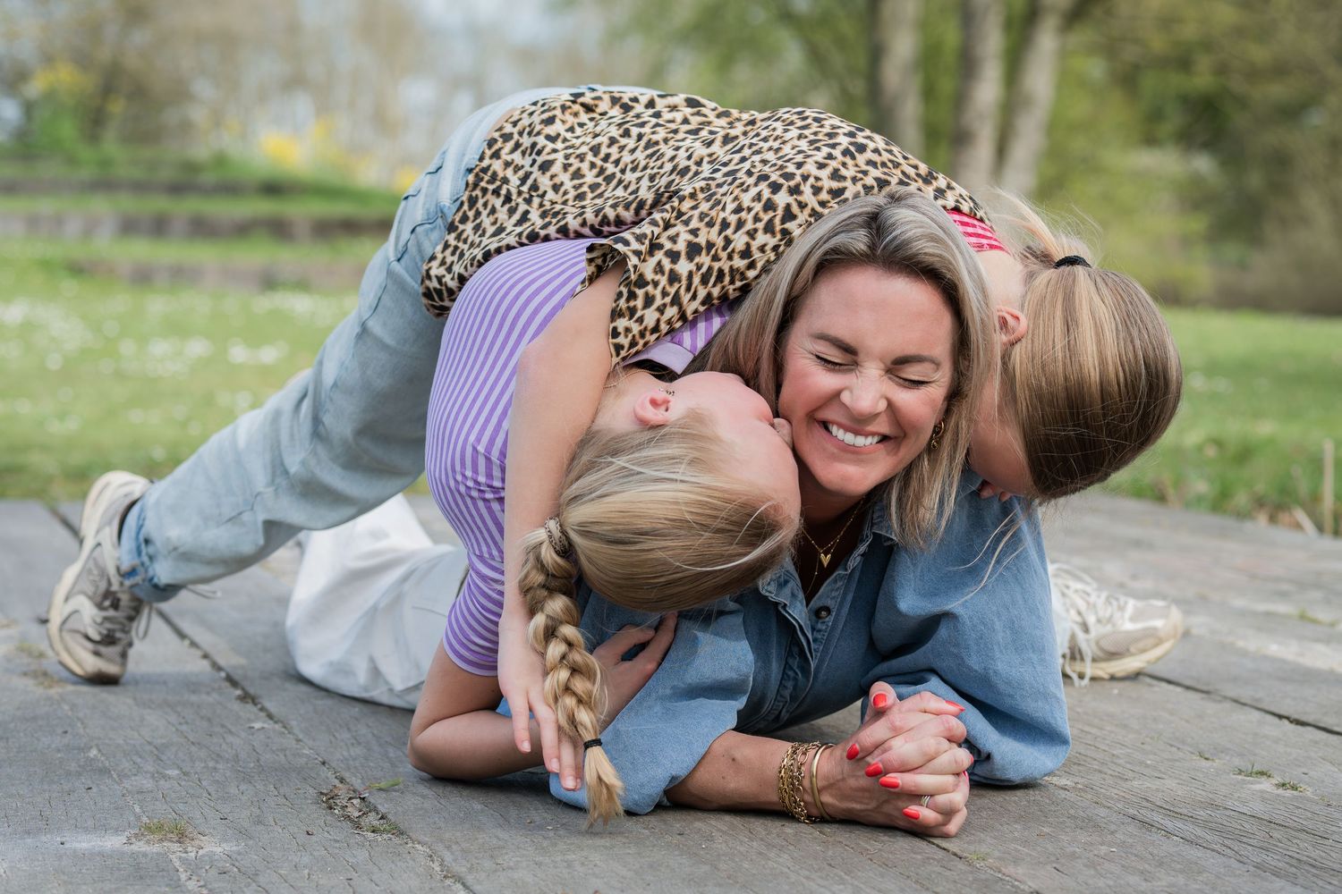 A smiling woman lies on the ground while two young girls hug and cuddle her outdoors in a park setting.