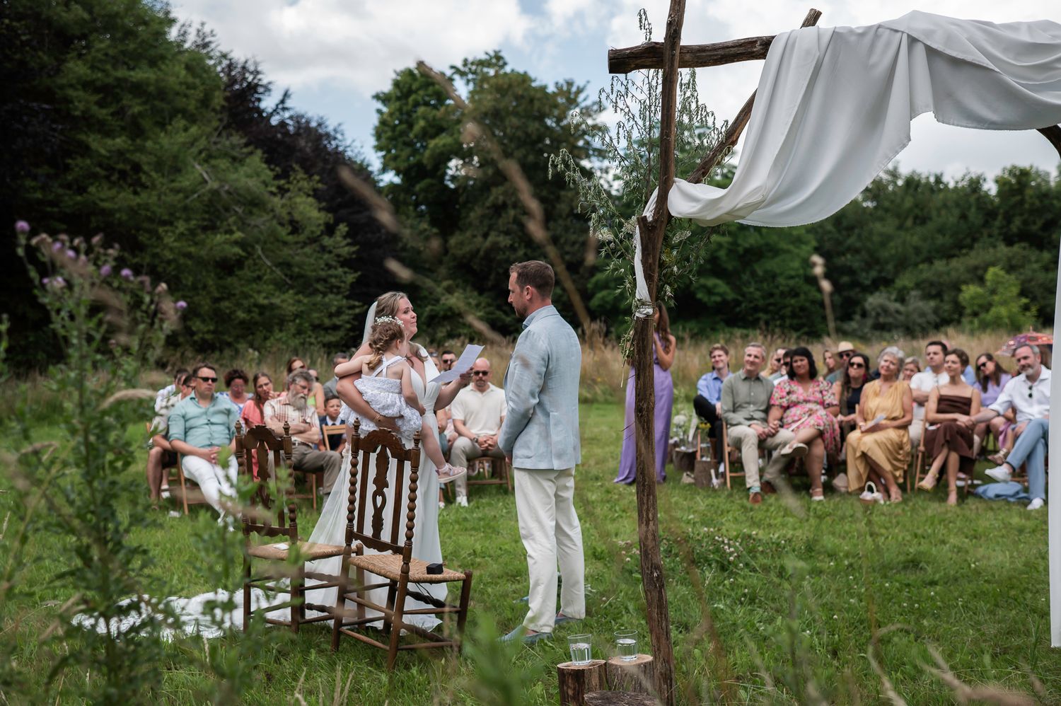 Outdoor wedding ceremony in a lush garden with guests seated, bride and groom standing at a rustic wooden cross altar.