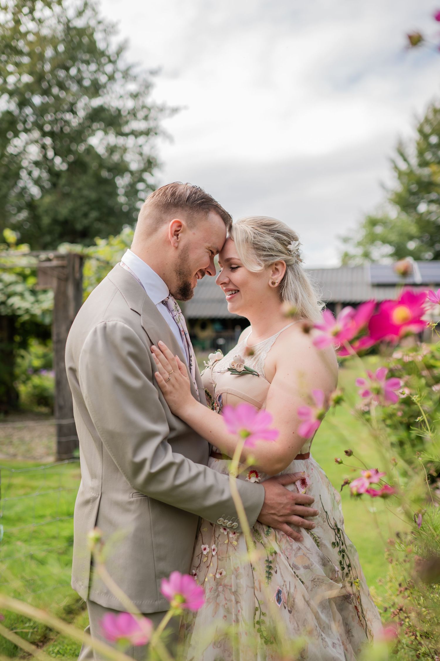 A couple shares a romantic forehead touch amidst pink cosmos flowers in a garden wedding portrait.