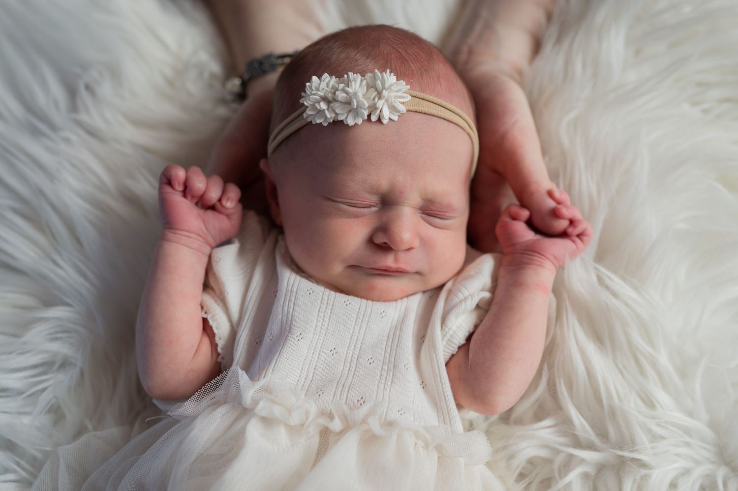 Peaceful newborn baby girl sleeping in white outfit and floral headband on fluffy white blanket.