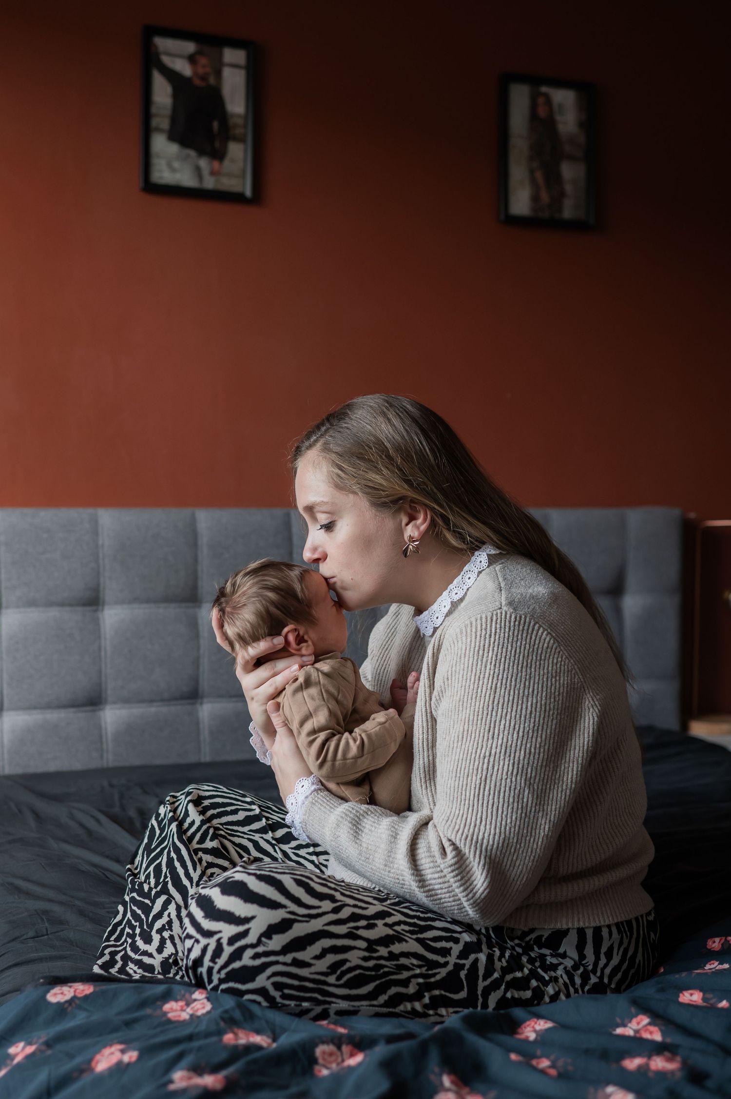 Young mother sitting on a bed kissing her newborn baby tenderly in a cozy red-walled bedroom.