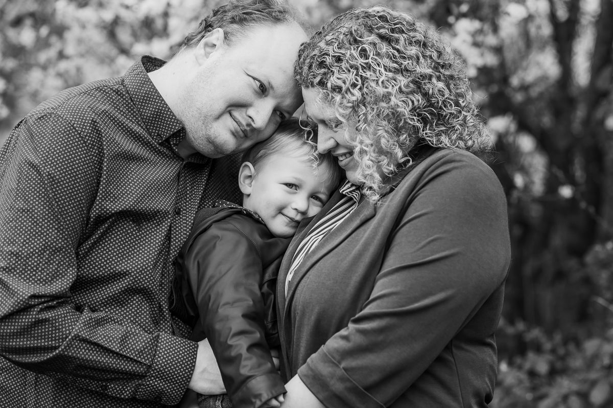 Black and white family portrait of parents lovingly embracing their young son outdoors among autumn foliage.