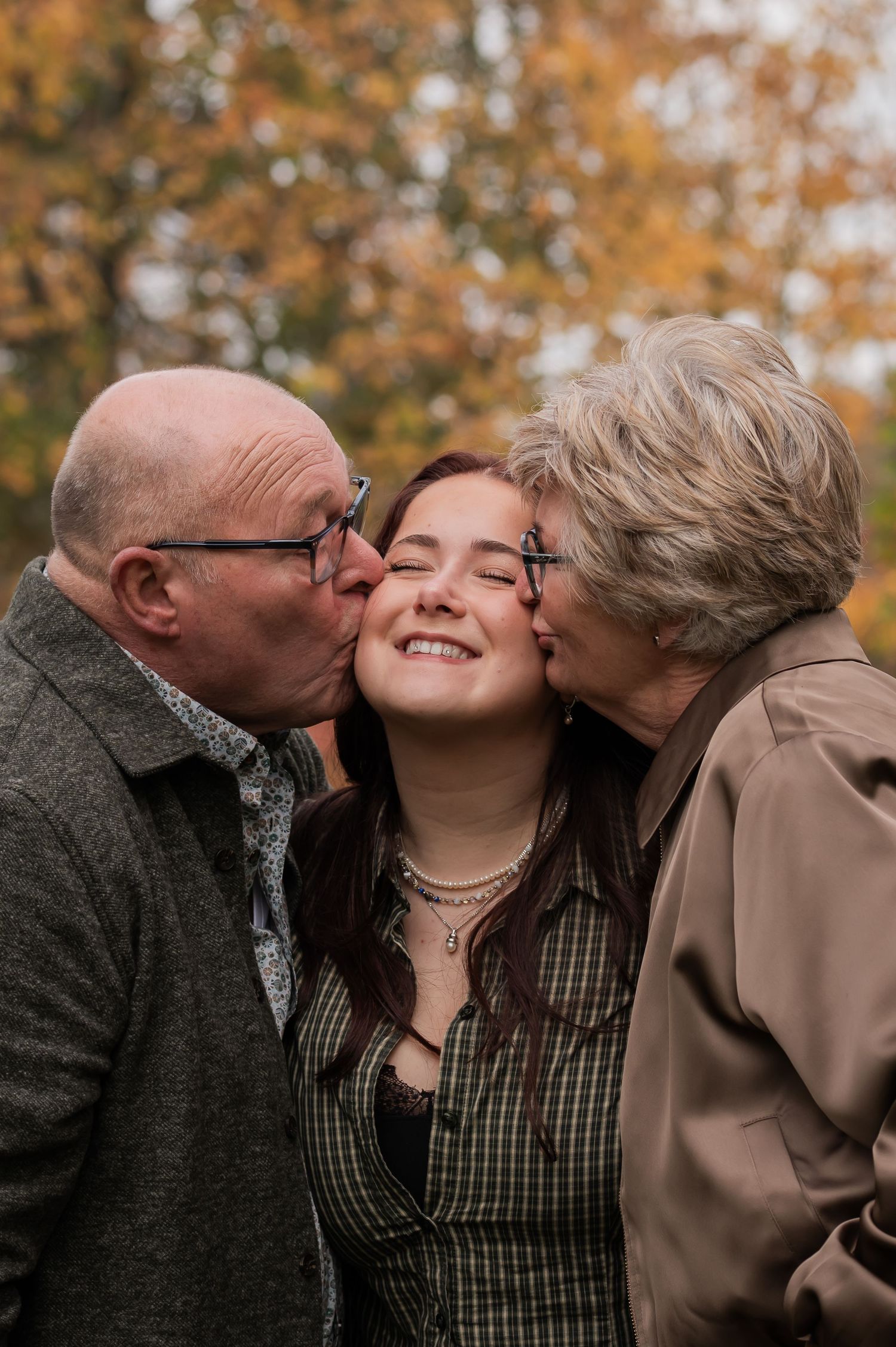 Elderly grandparents kissing their smiling granddaughter on cheeks outdoors in autumn park.