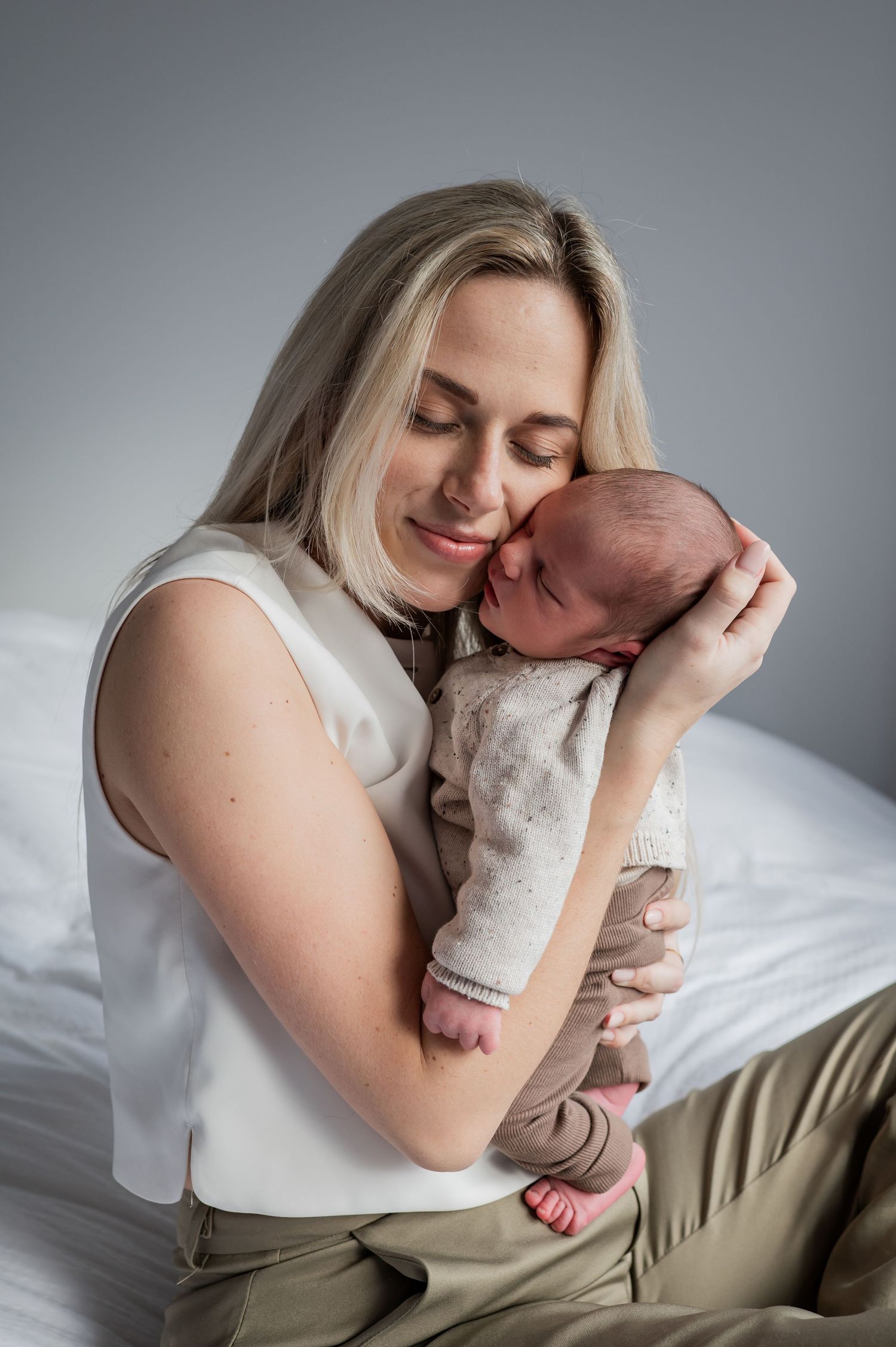 Blonde woman in white top gently pressing cheek against sleeping newborn baby held in her arms.