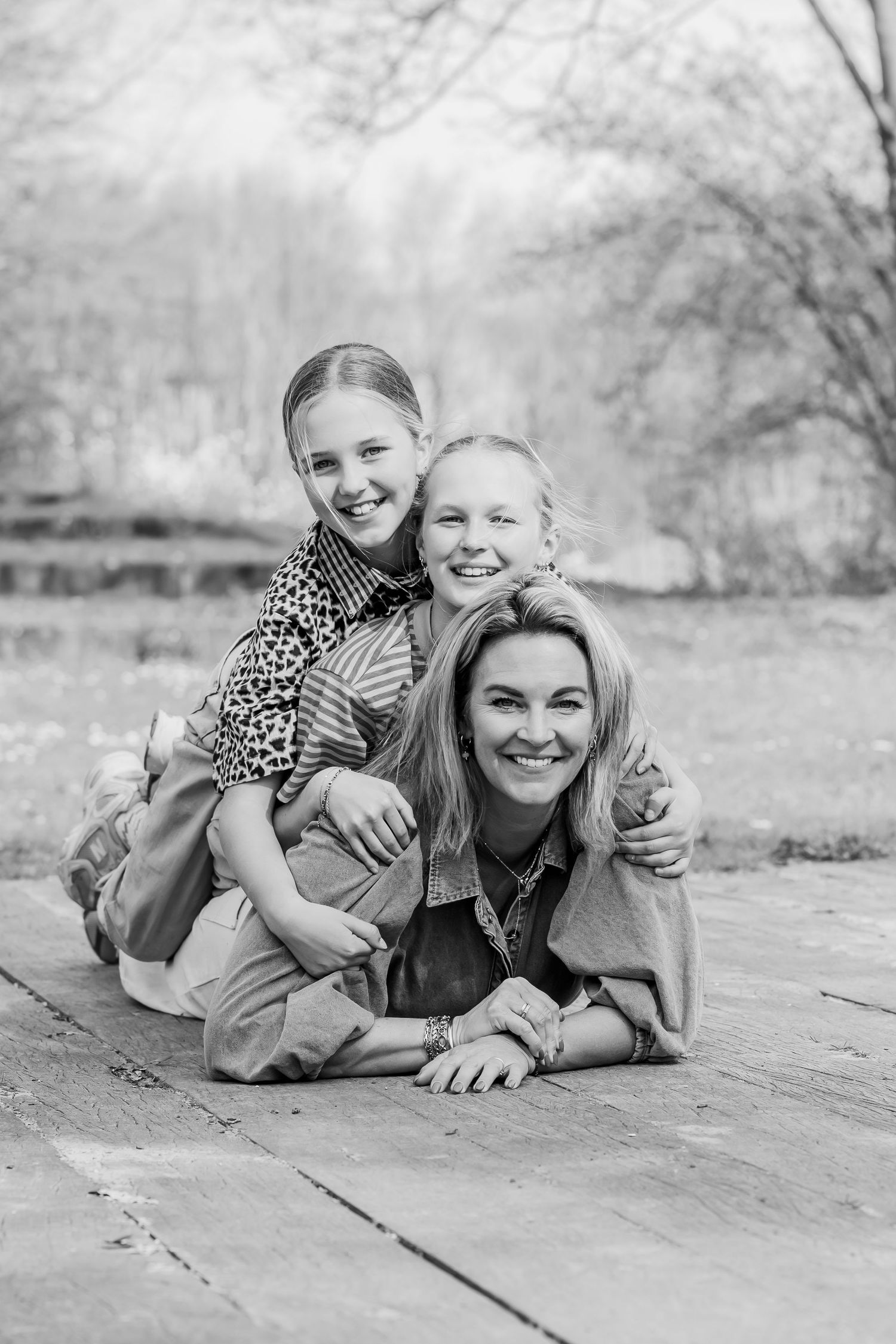 Black and white photo of a smiling mother lying on wooden deck with two happy daughters sitting on her back outdoors.