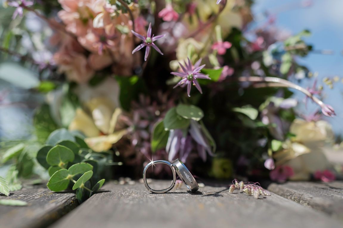 Two wedding rings resting on a rustic wooden surface with a soft floral bouquet in the background.