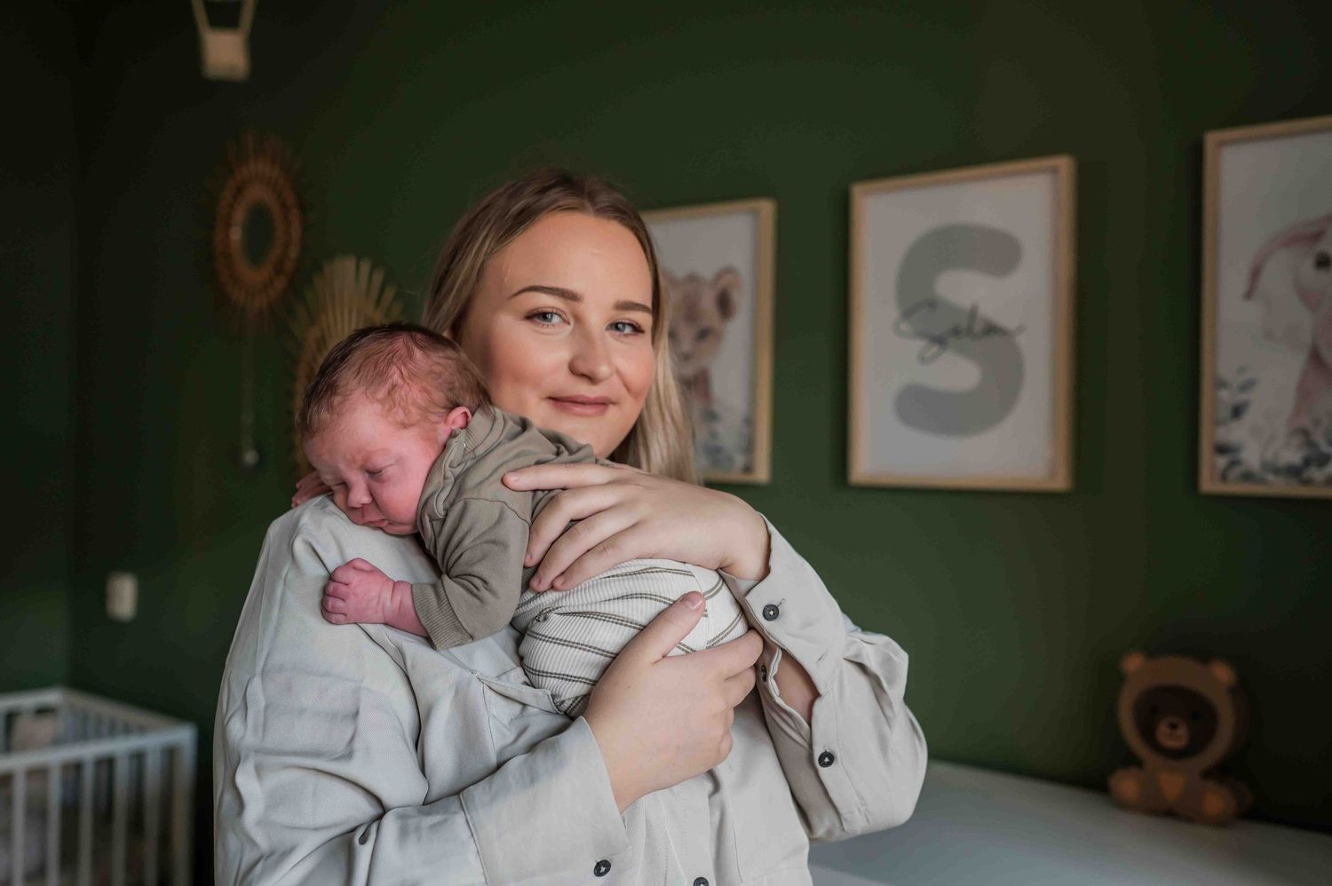 Smiling young mother holding her sleeping newborn baby in a green nursery room.