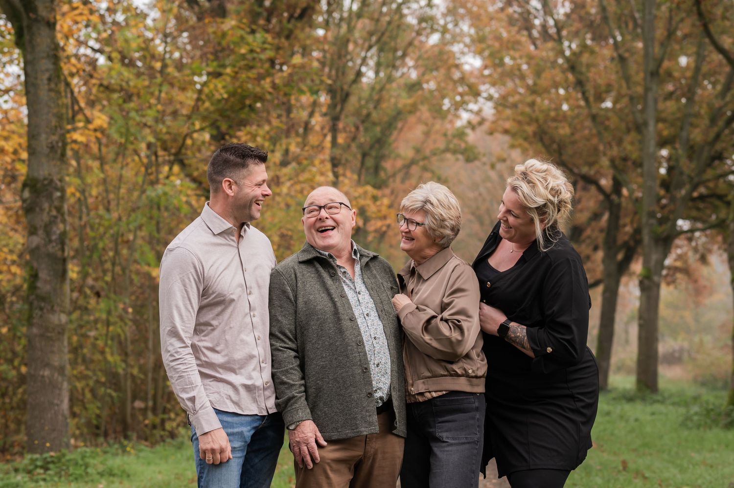 Four adults laughing together outdoors in an autumn forest with colorful fall foliage.