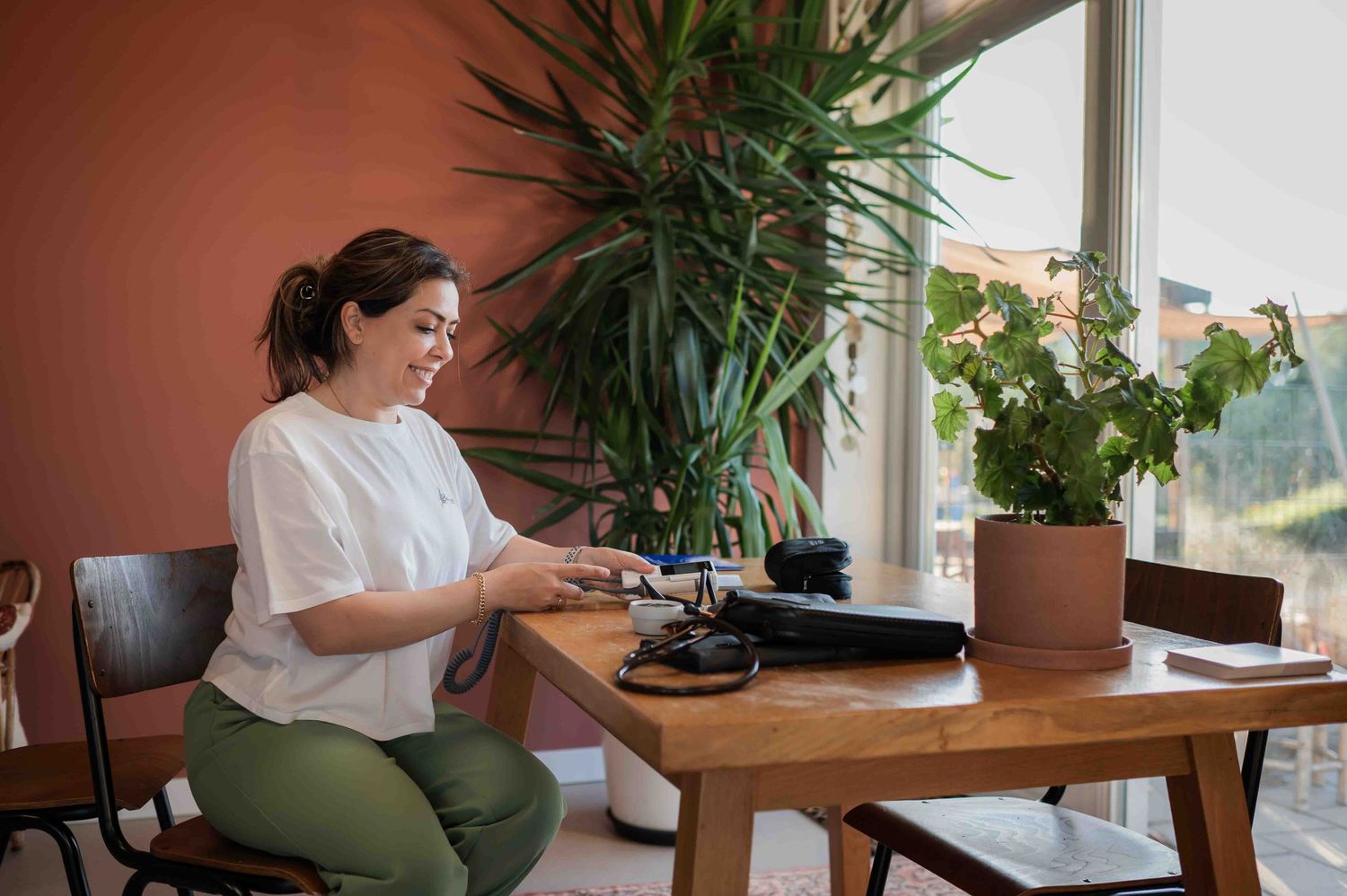Woman in white blouse sitting at wooden desk with plants, using a device near a bright window indoors.