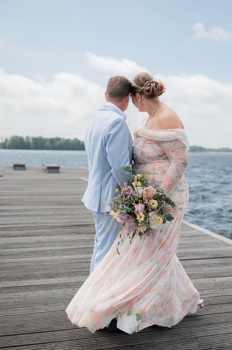 Bride in floral off-shoulder gown holding bouquet, touching foreheads with groom on wooden dock by lake.