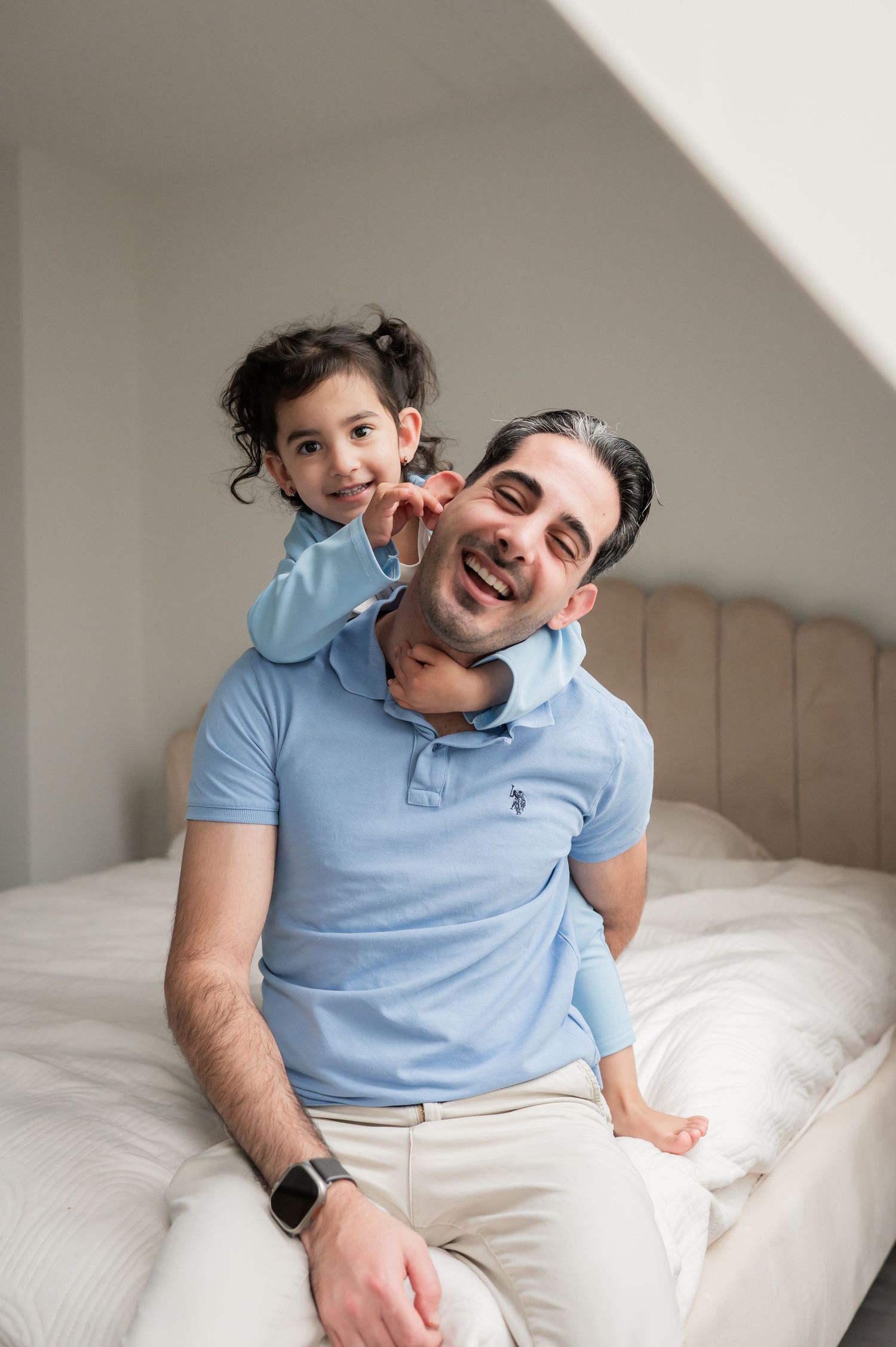 Laughing father in blue polo shirt with curly-haired young daughter playfully perched on his shoulders on bed.