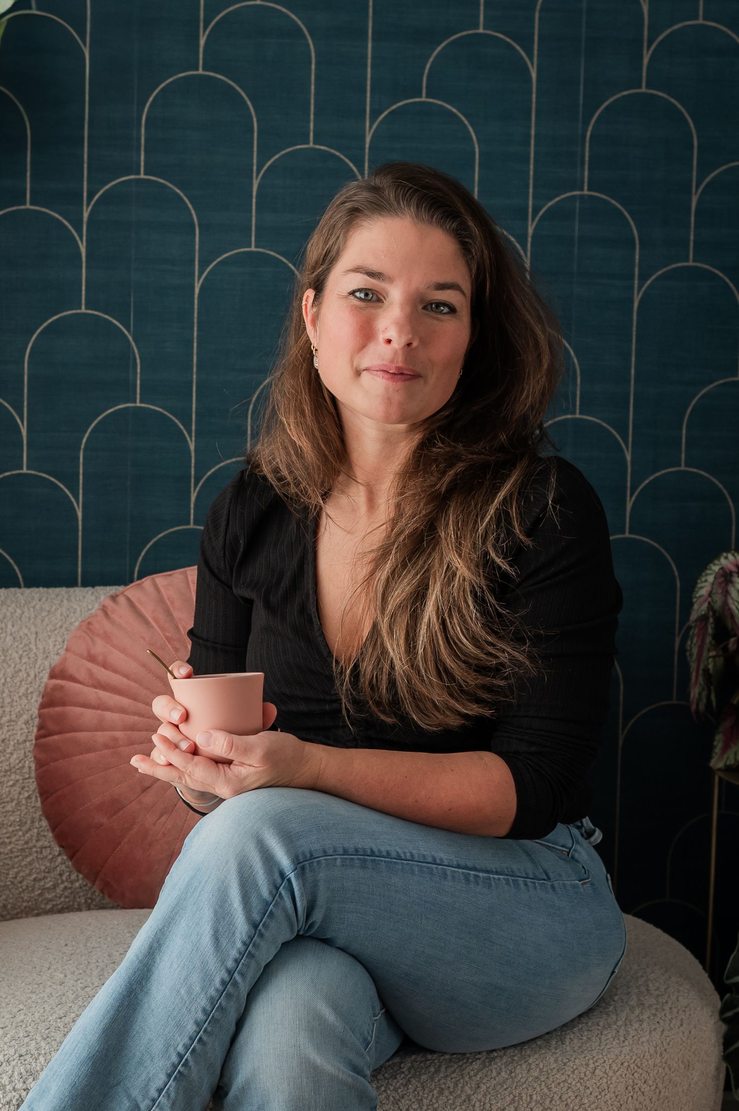 Woman sitting relaxed on white chair holding pink mug, wearing black top and jeans against teal arch wallpaper.