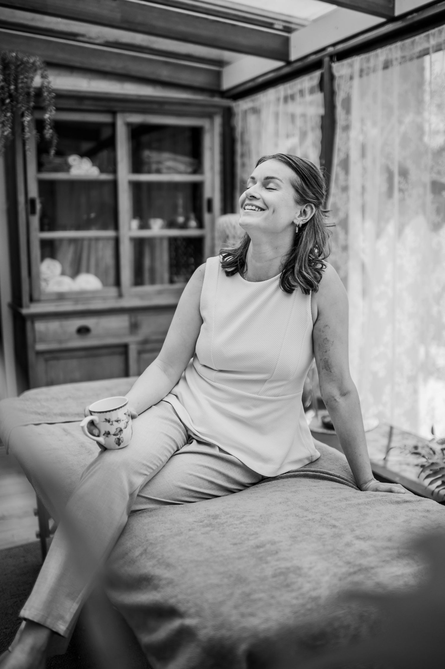 A smiling woman sits on a bed holding a mug, dressed in a sleeveless outfit, in a cozy black and white interior photo.