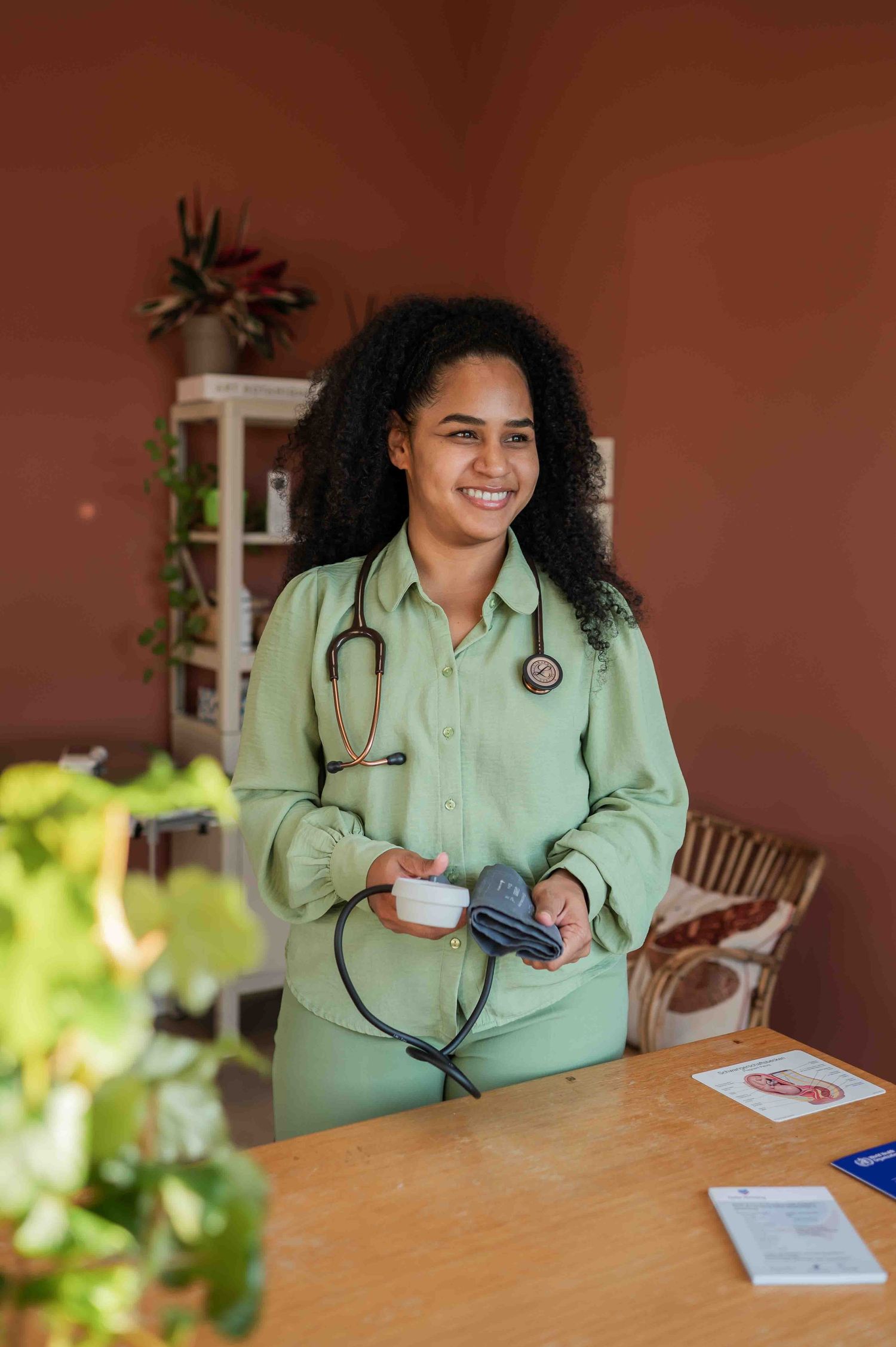 Smiling female nurse in green scrubs holding a blood pressure monitor with a stethoscope around her neck.