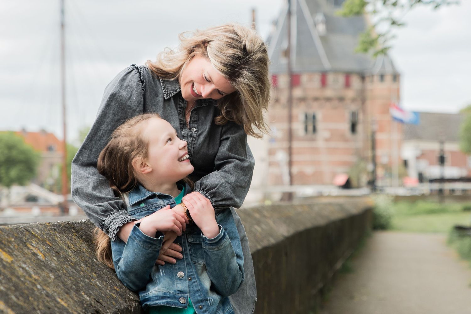 A mother and young daughter share a joyful moment leaning on a stone wall with a historic Dutch building in the background.